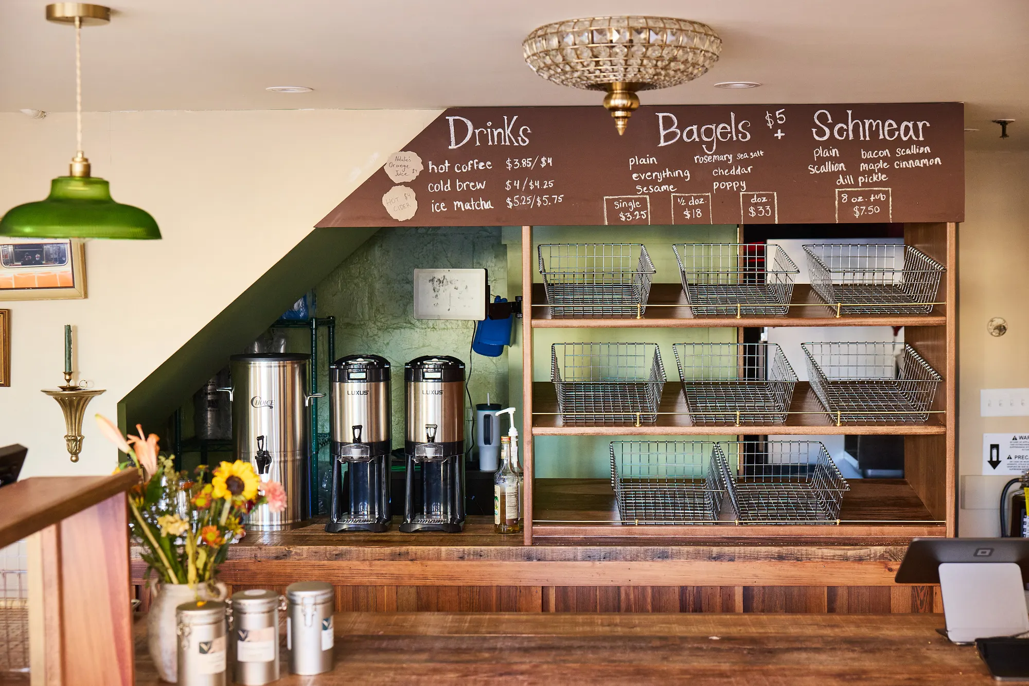 The menu wall at Brick Street Bagels, a chalk board over wire baskets and coffee urns, under a crystal light