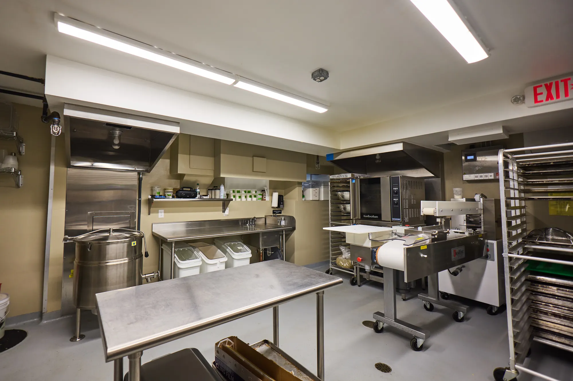 The bakery behind the shop: steel tables, a kettle, ovens and sheet-pan racks under fluorescent light.