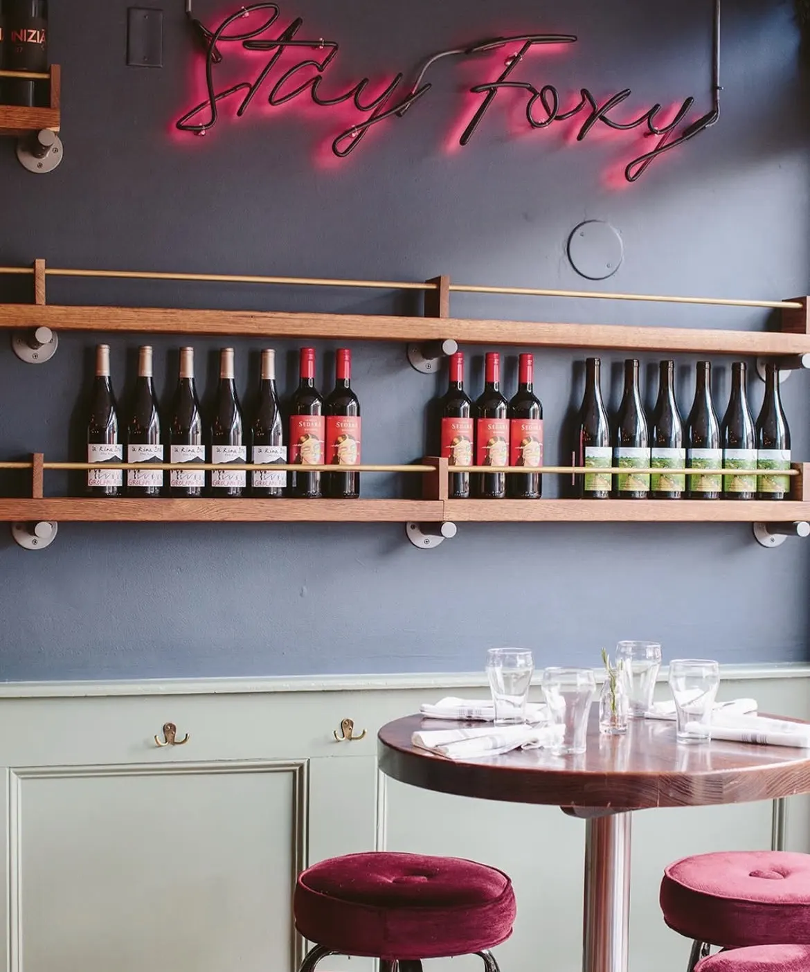 A pink neon sign reading Stay Foxy above timber shelves of wine, with a high table and velvet stools below.