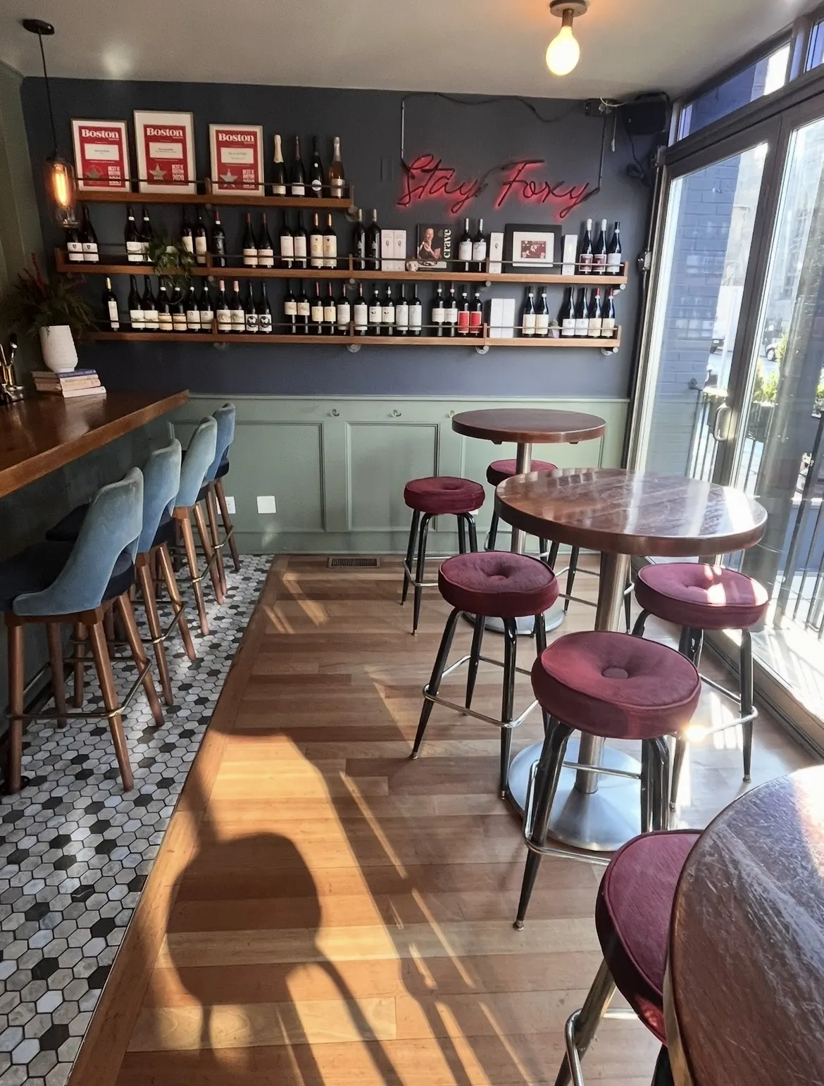 Burgundy velvet stools at round high tables beside the glazed front, under the Stay Foxy sign and a wall of wine.