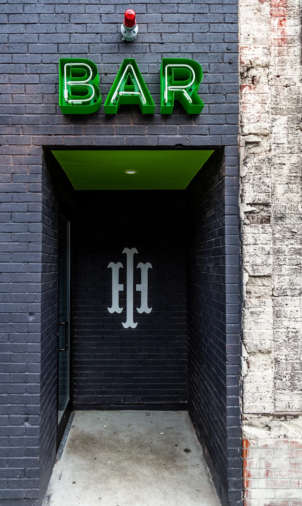 A green neon BAR sign over a dark brick doorway marked with a monogram.