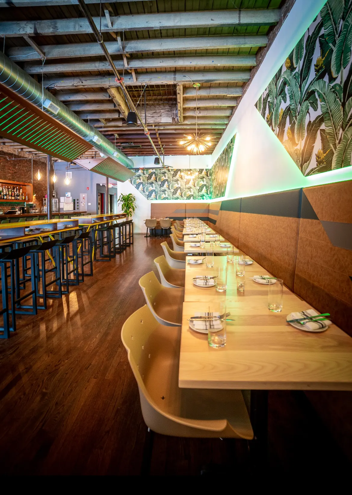 Long tables under a palm-print mural and green-lit beams.