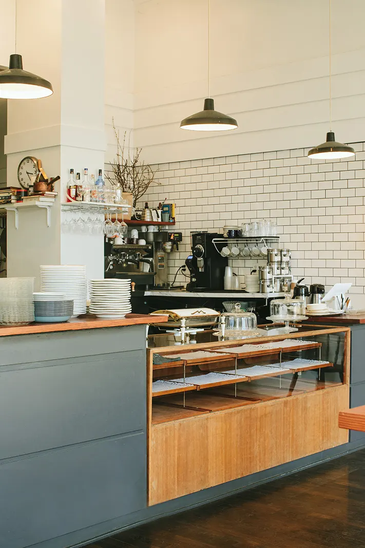 A pastry case and espresso machine at a counter against white subway tile.
