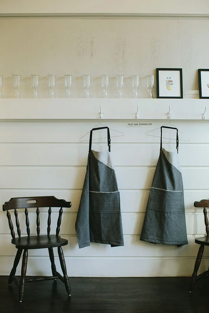 Two aprons hung on hooks on a white-panelled wall, beside a timber chair.