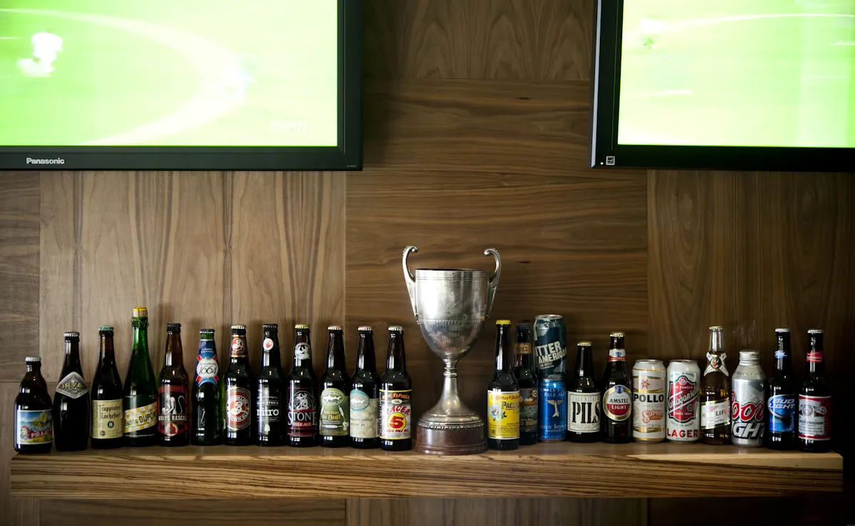 Beer bottles and a silver trophy on a shelf below two televisions.
