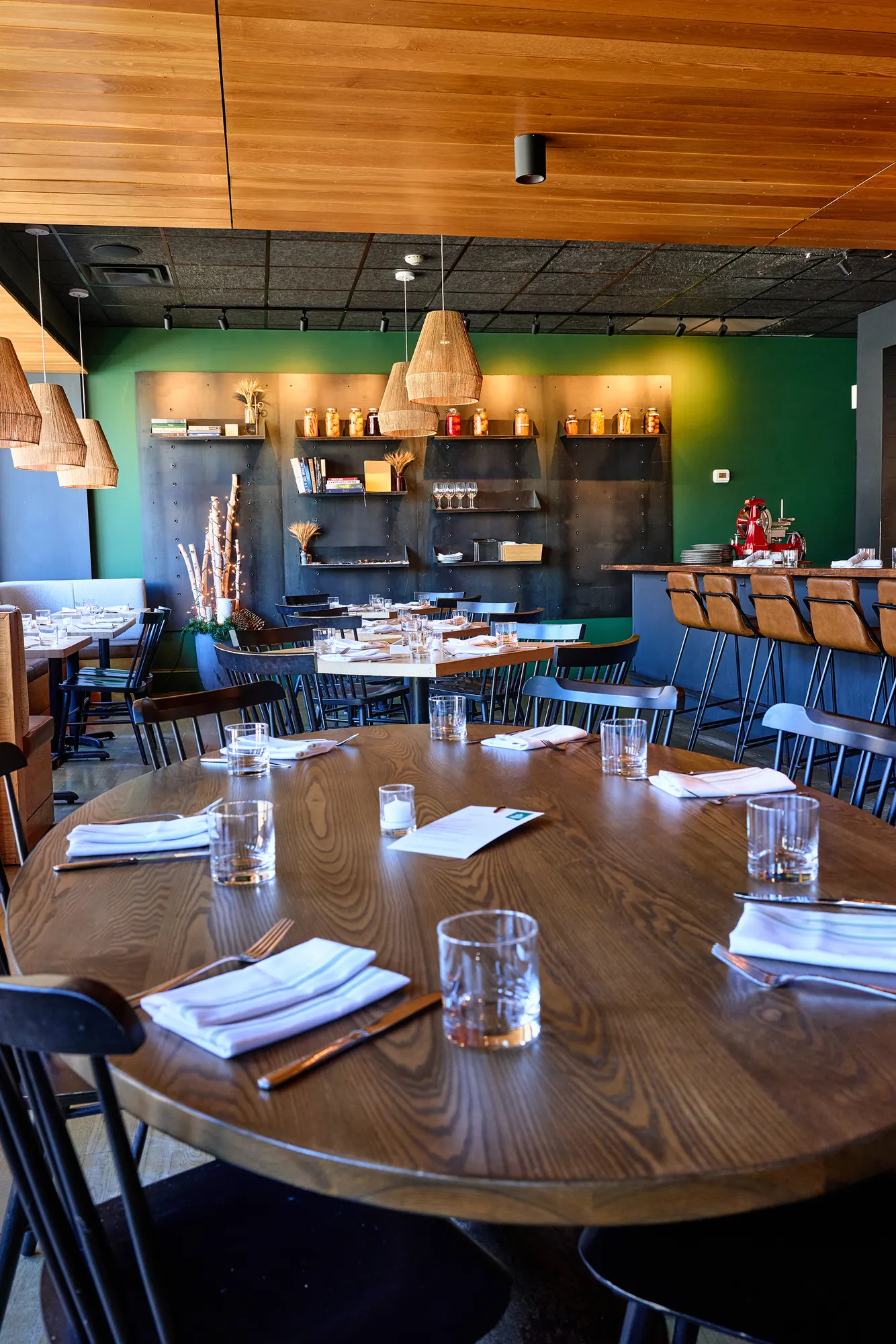 A round walnut table set for dinner, with the green shelving wall and woven pendants beyond.