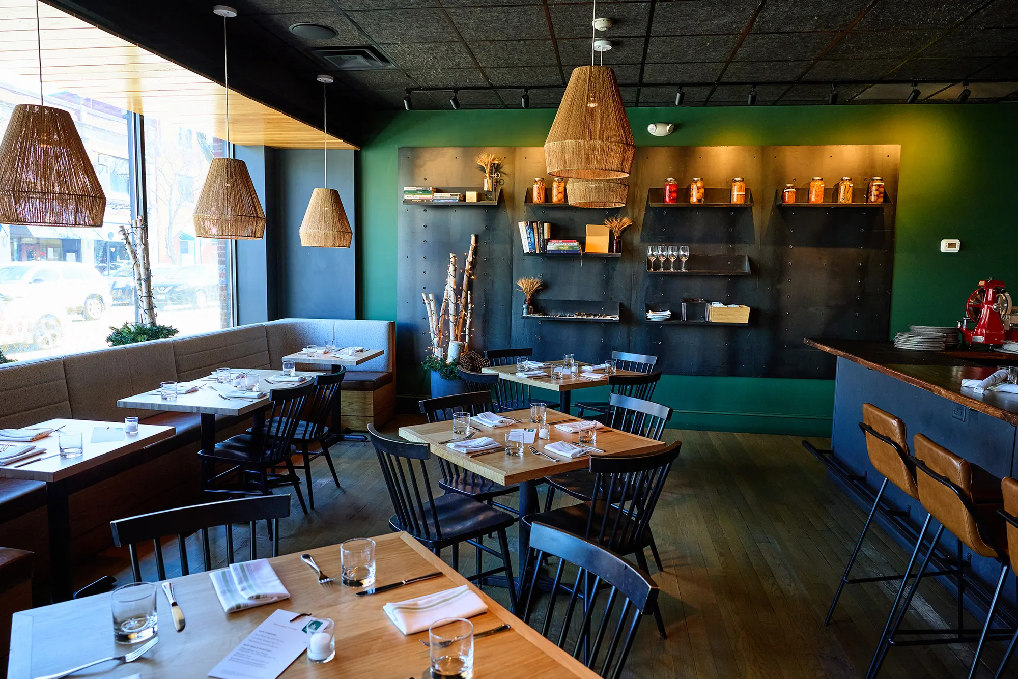 Tables and black chairs under woven pendants, with the green shelving wall at the back.