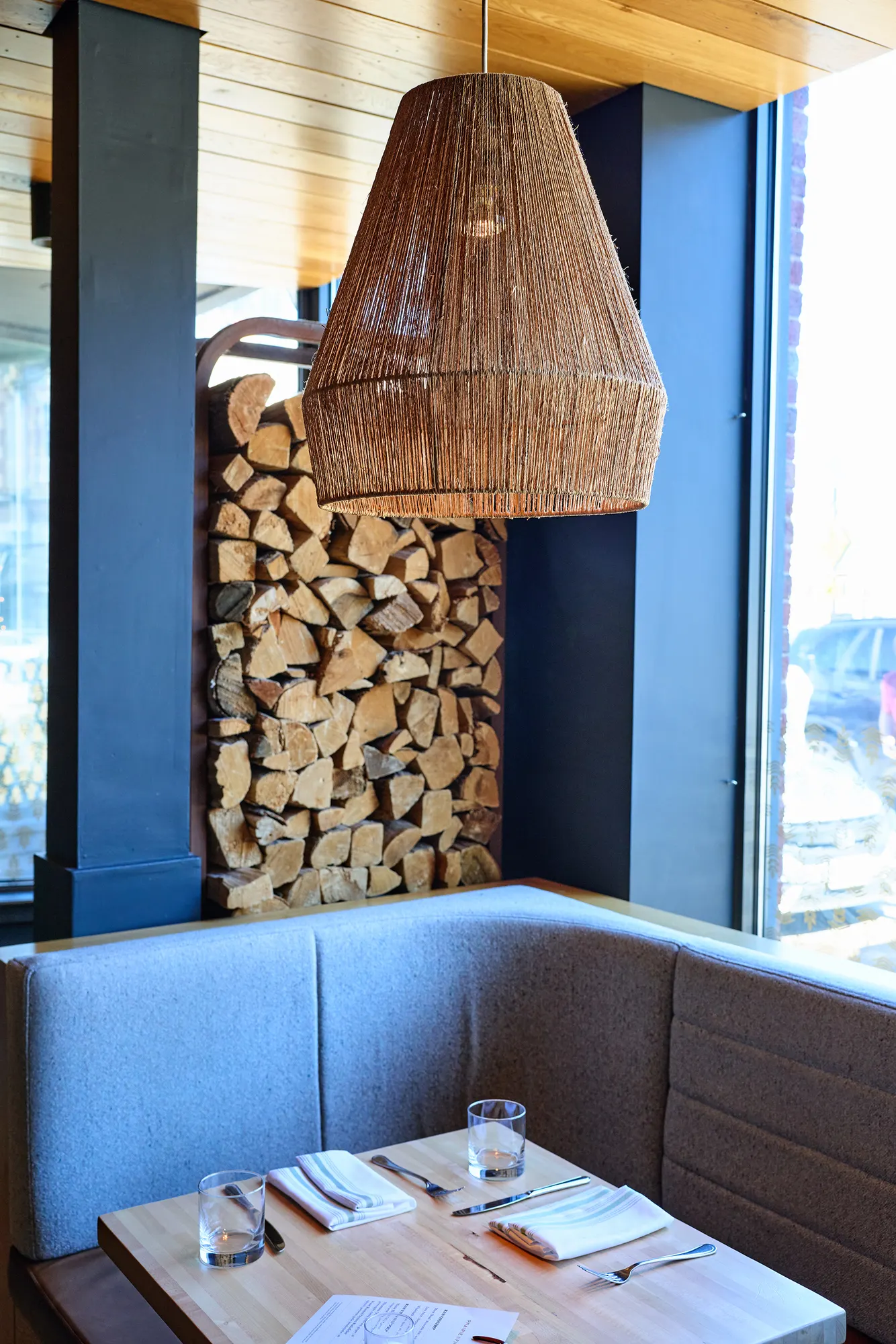 A banquette table beside a column of stacked firewood, under a woven pendant.