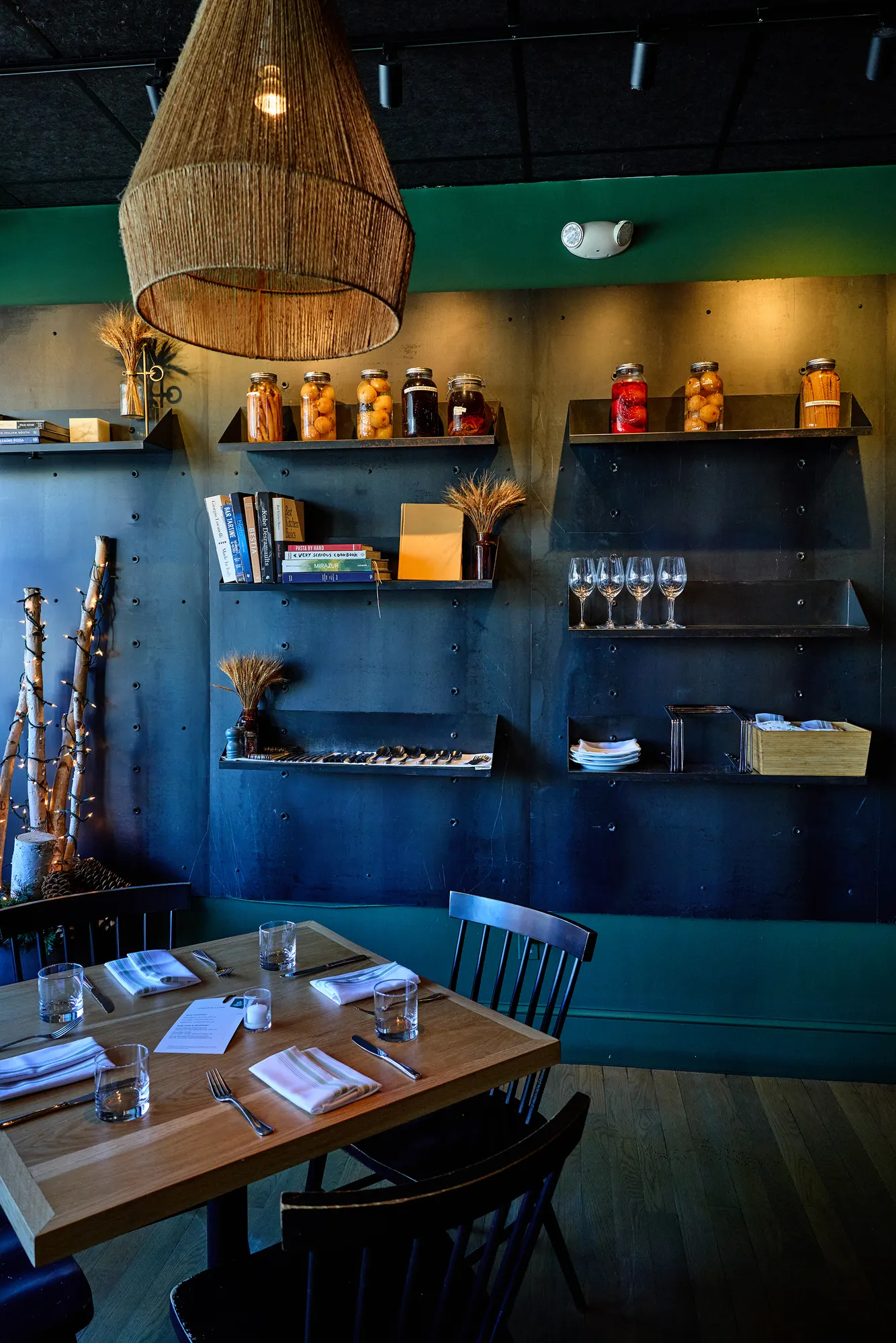 Open steel shelves of jars, books and glassware against the green wall, over set tables.