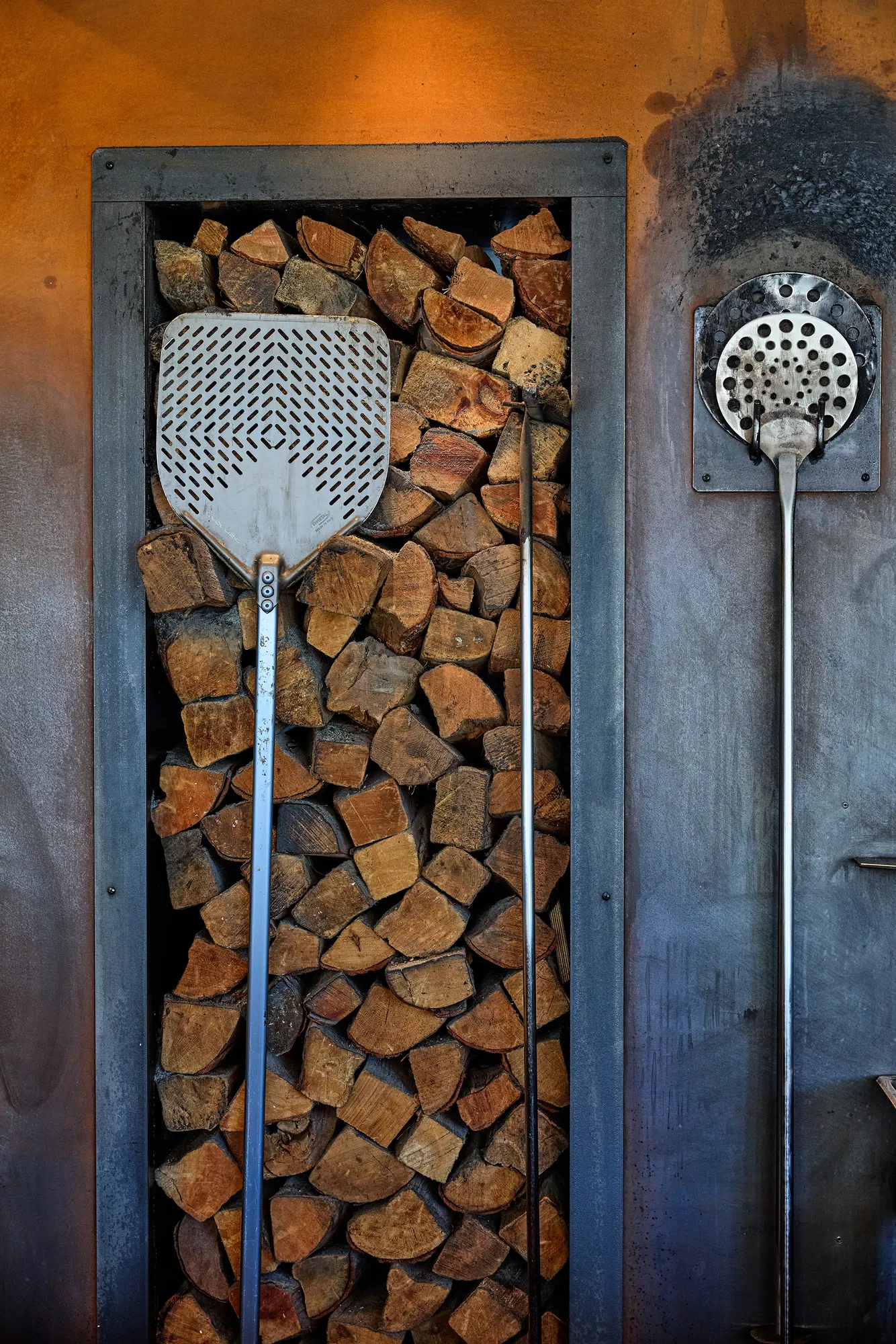 A pizza peel and an oven brush hung against a niche of stacked firewood.