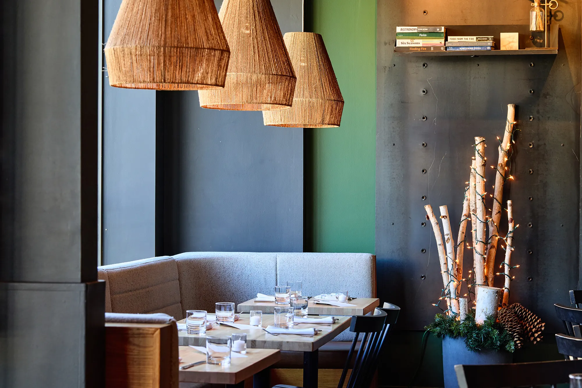 The grey banquette and window tables under woven pendants, with birch trunks in the corner.