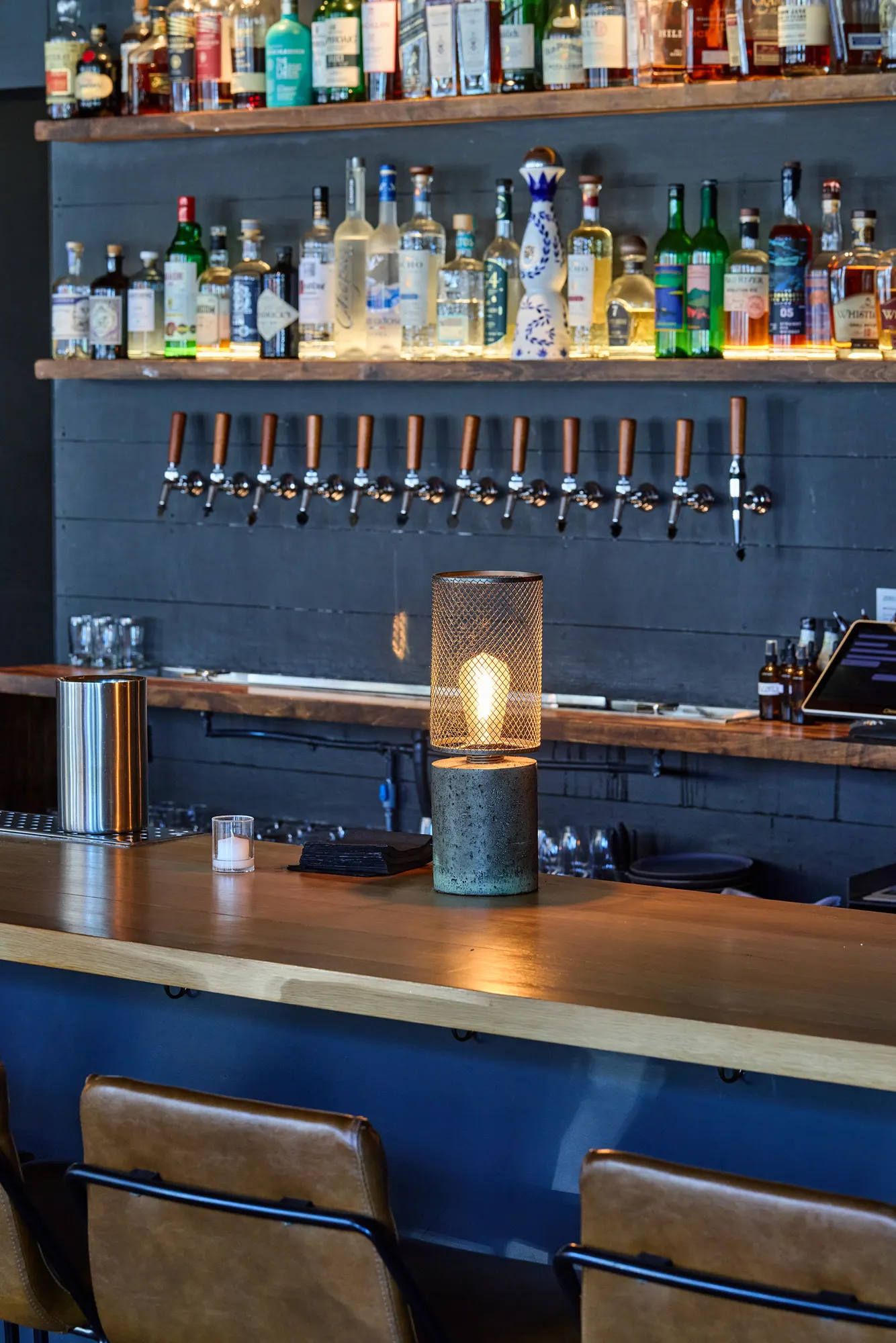 Bottles on lit shelves over a row of taps, with a lantern on the bar top.