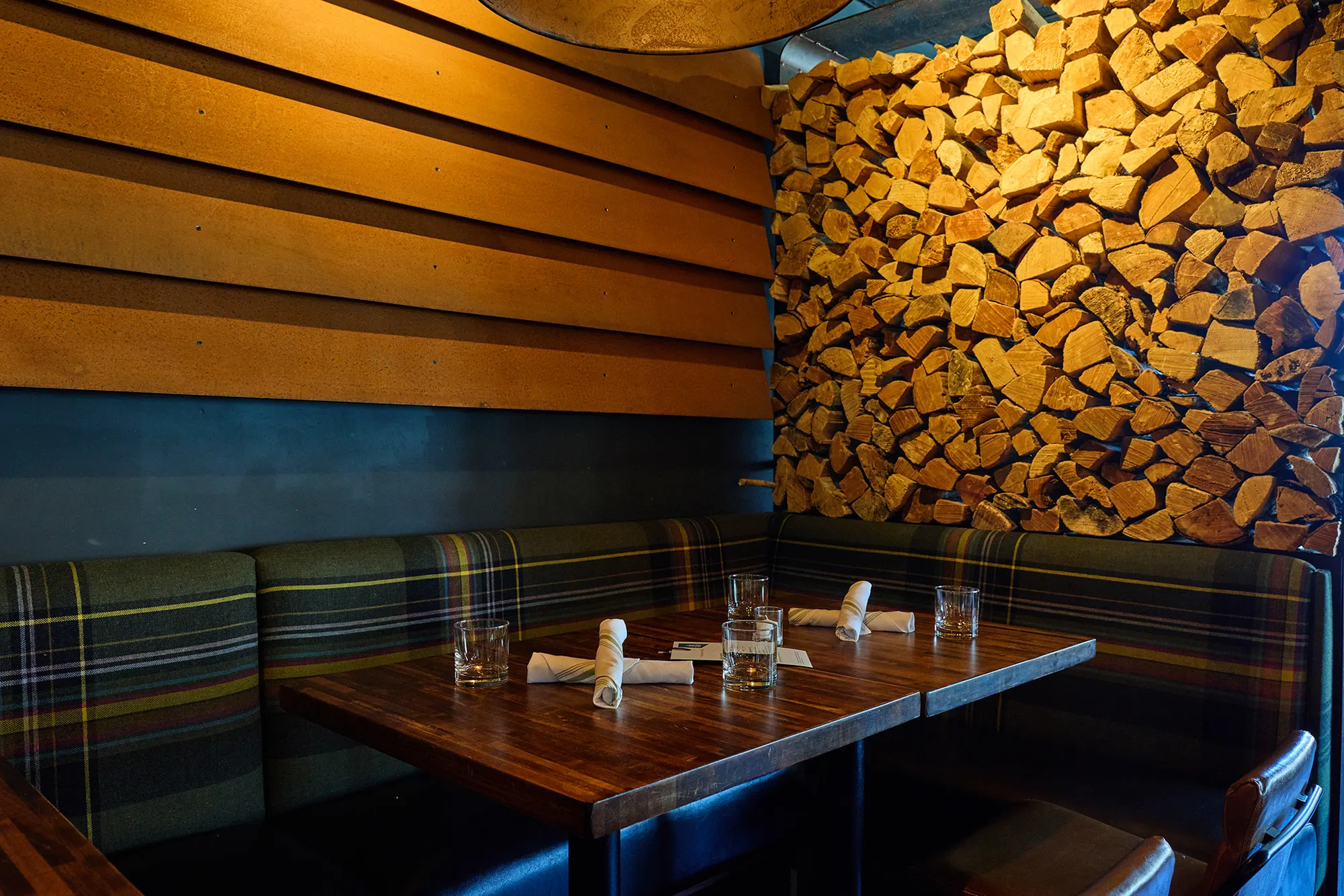 The plaid banquette meeting the firewood wall below the timber boards.