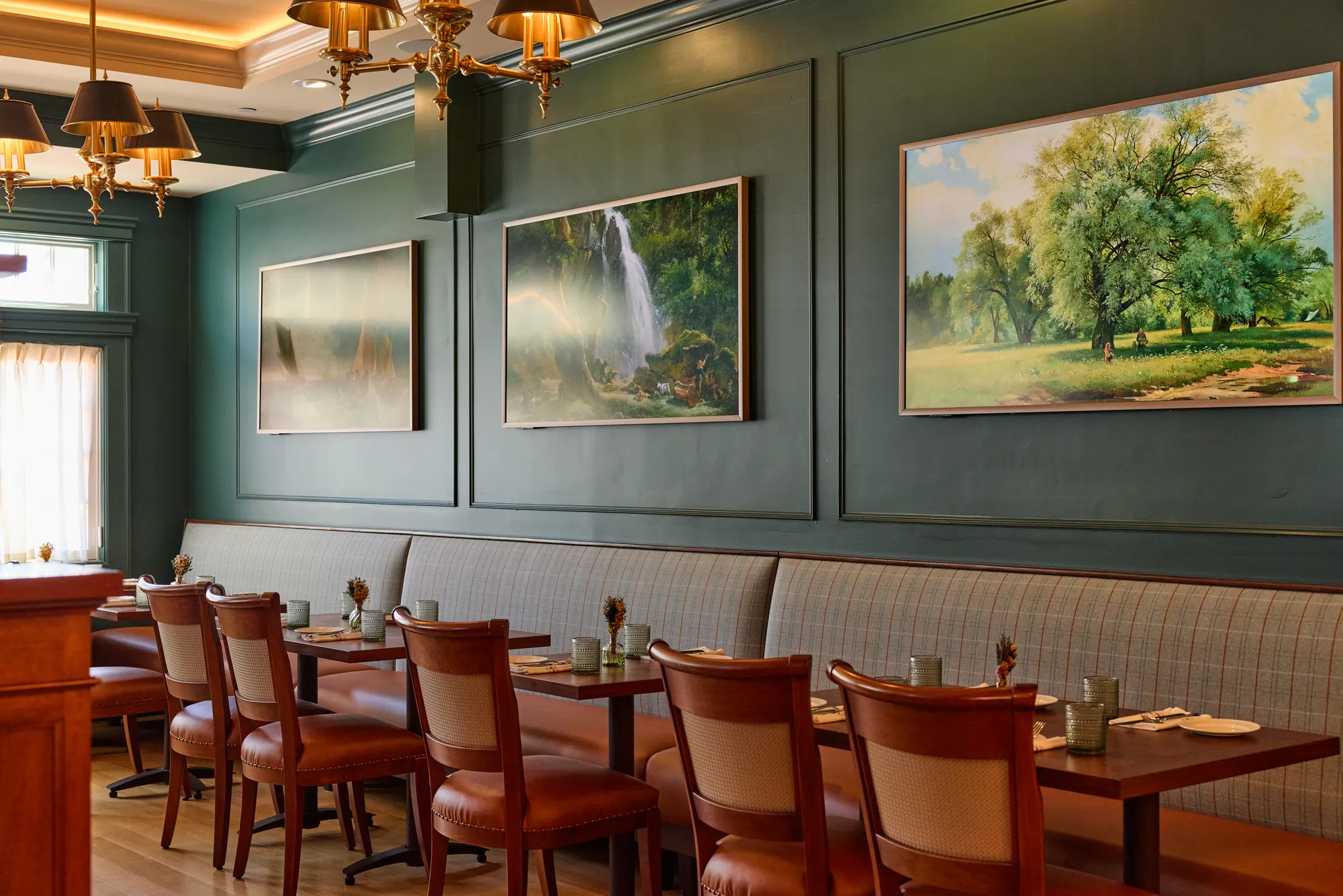 Landscape paintings on deep green panelling above a long plaid banquette, with timber tables and leather-seated chairs under brass chandeliers.