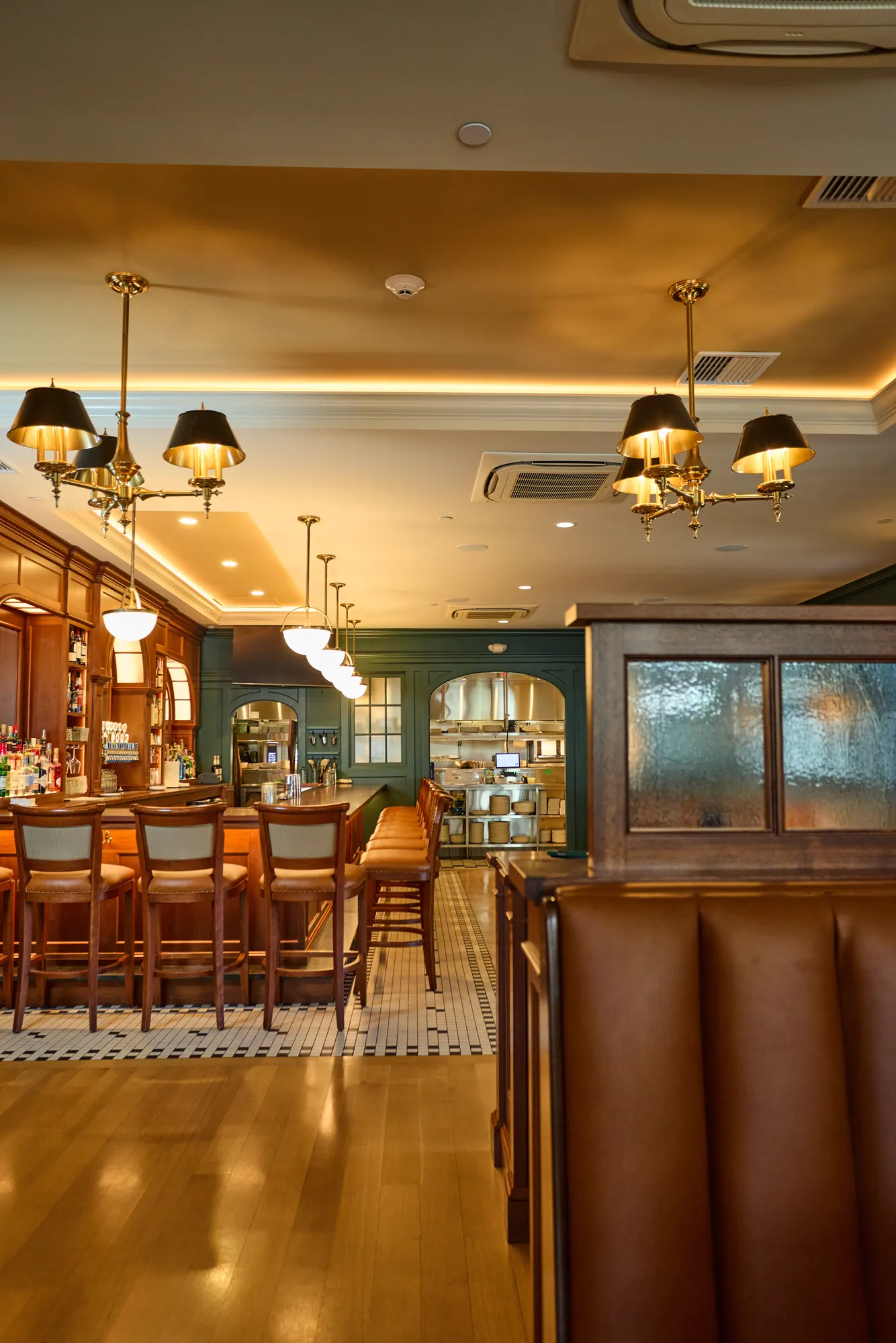 Bar stools along a timber bar under brass pendants with black shades, a black-and-white mosaic floor at its foot, and a leather booth in the foreground.