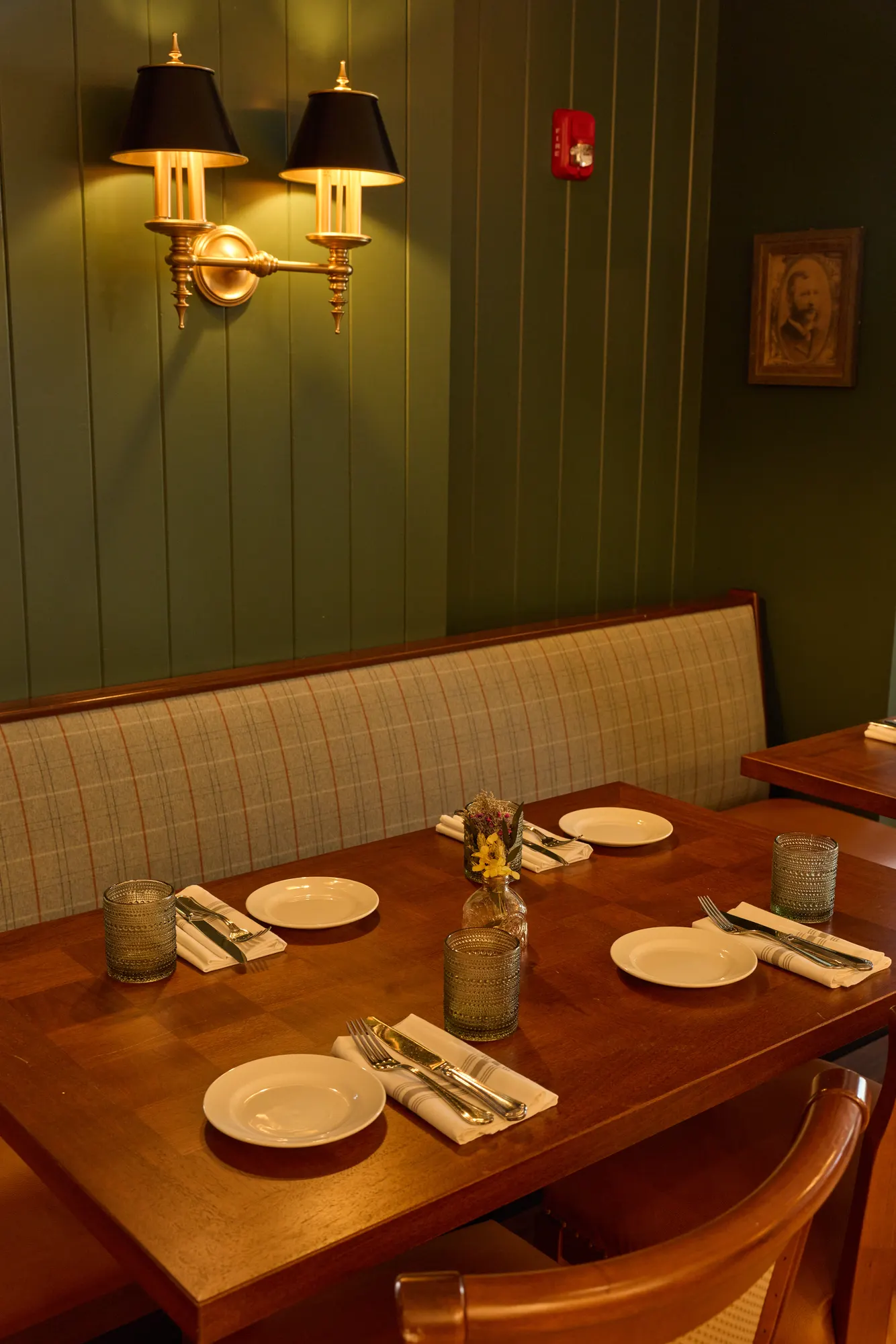 Plates and folded napkins on a timber table against a plaid banquette, below a brass double sconce with black shades on green board panelling.
