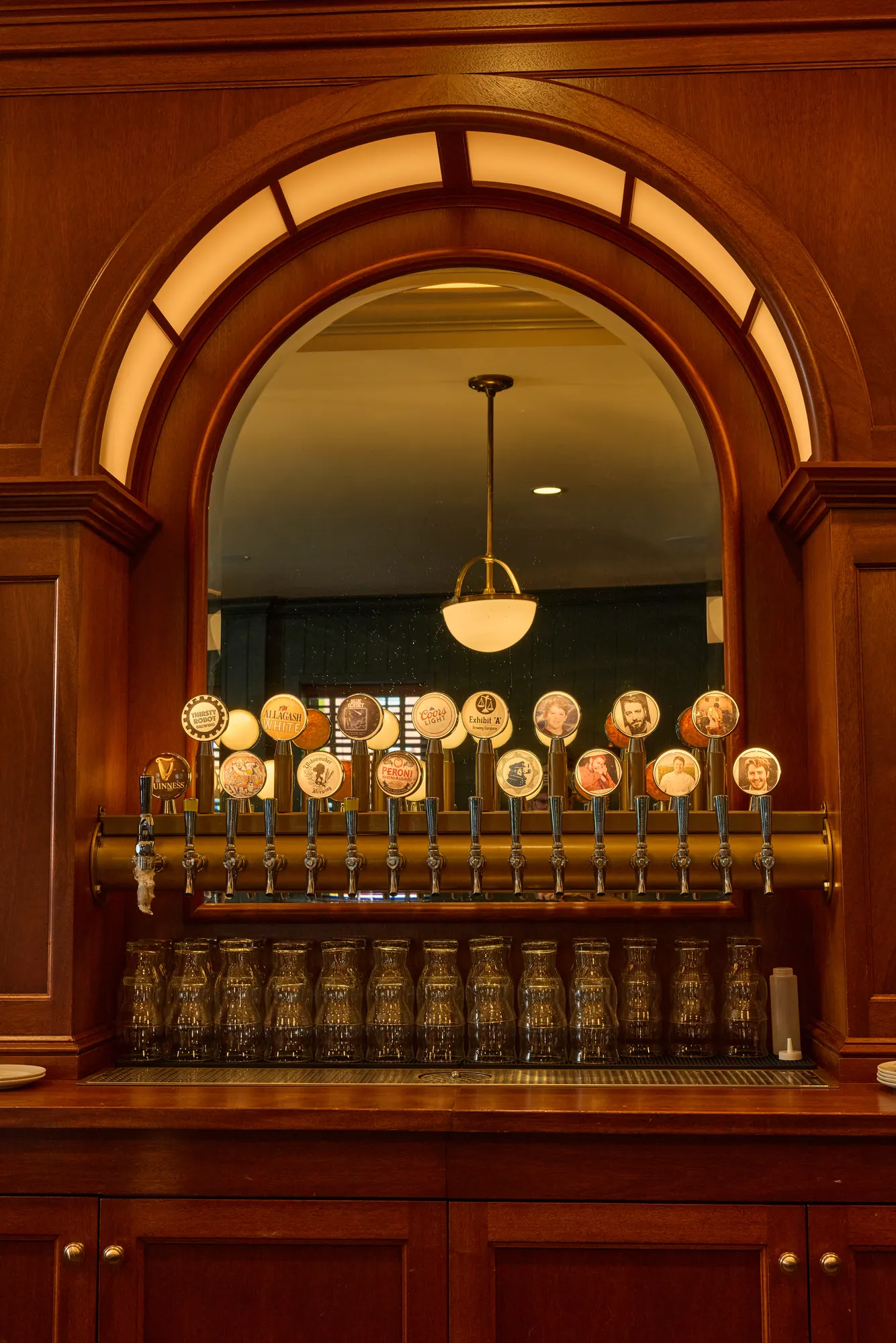 A row of brass beer taps with round badges, glasses lined up beneath, inside a mahogany arch with a lit fanlight around a mirror.