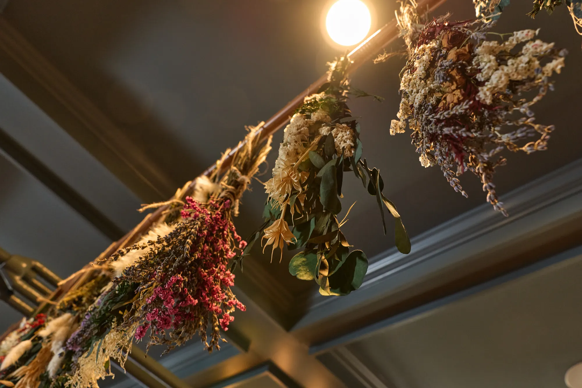 Bundles of dried flowers hanging upside down from a rail against the dark ceiling, beside a bare bulb.