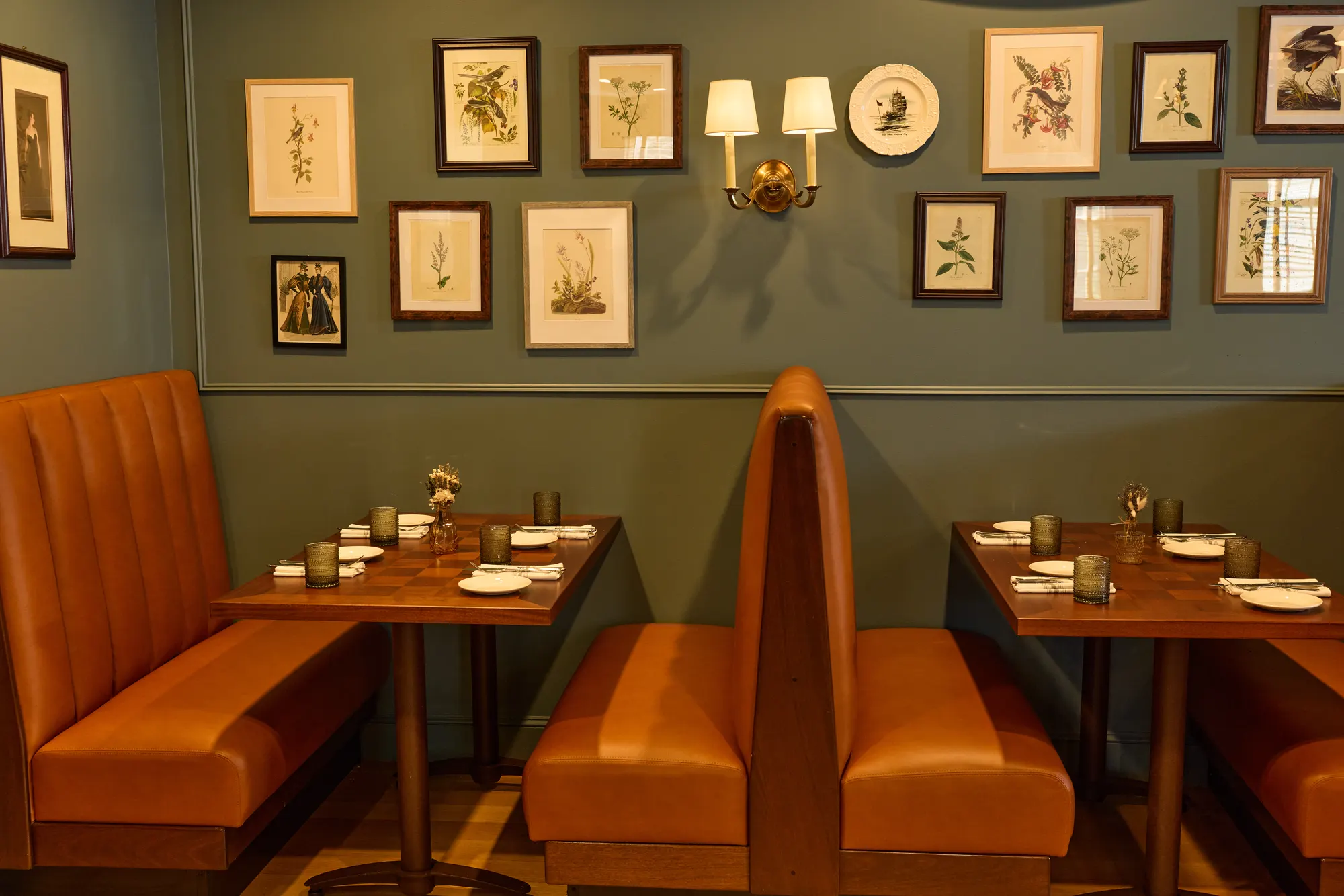 Tan leather booths with set tables beneath a wall of framed botanical prints and plates, a brass sconce among them, on blue-green paint.