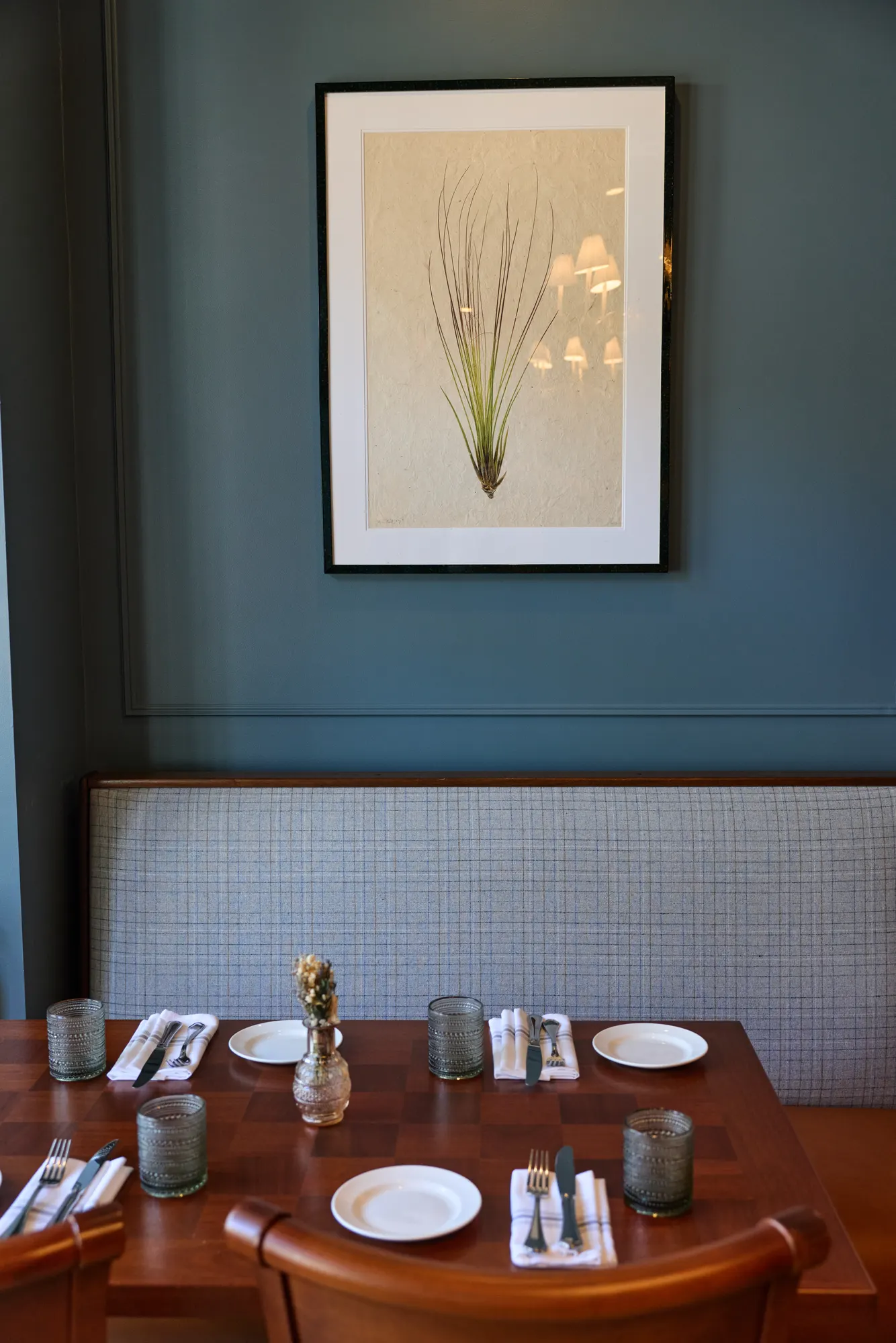 A framed print of a tall grass on a blue wall, above a plaid banquette and a set table.