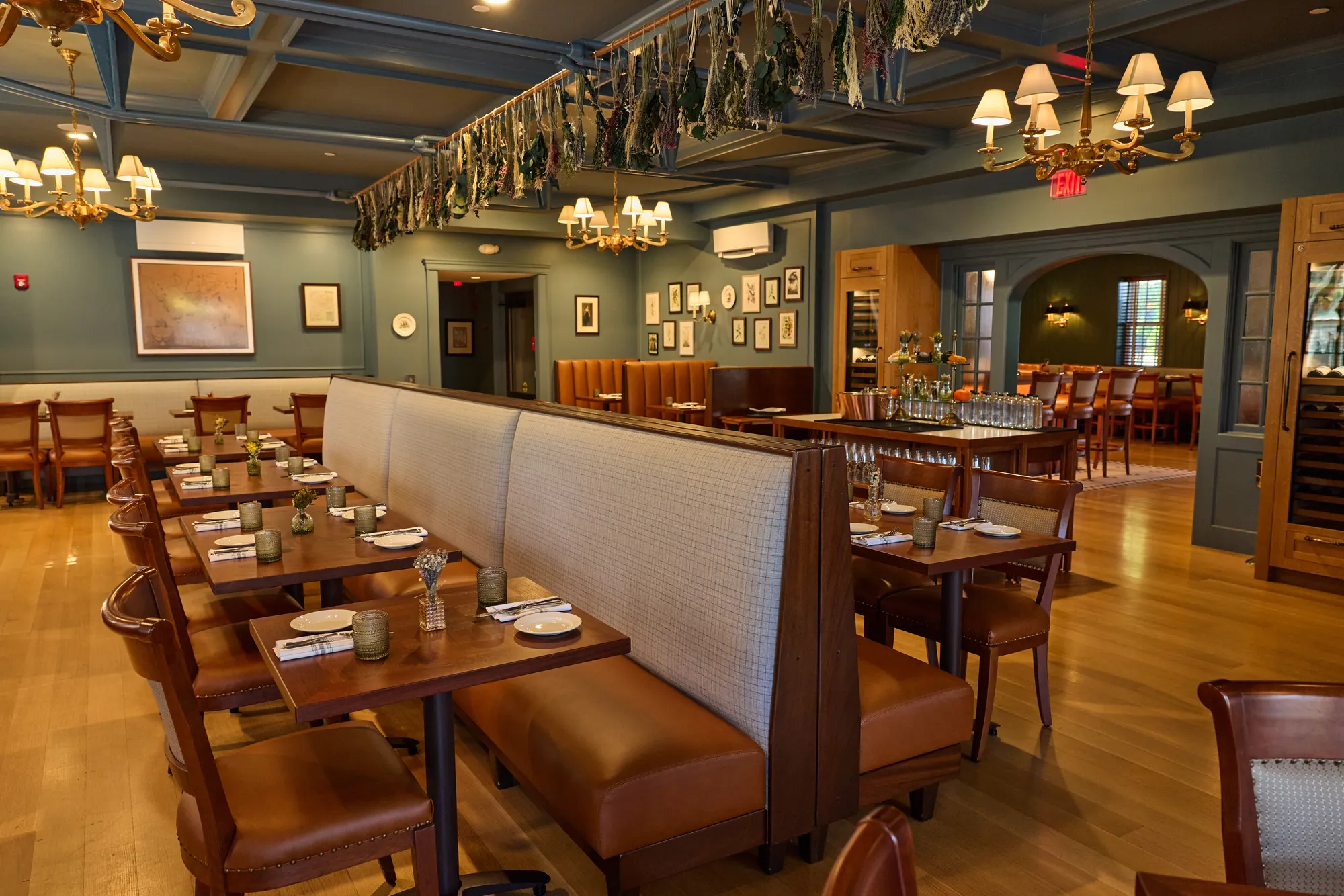 A high plaid-backed banquette dividing two rows of tables under the coffered ceiling and its dried flowers, with an arched doorway at the far end.