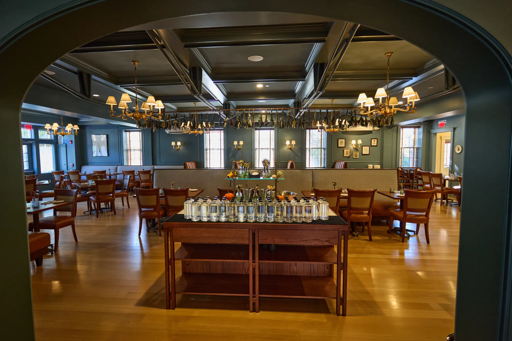An arched opening framing the dining room, a serving table of glassware at its centre, under a coffered ceiling hung with dried flowers and chandeliers.