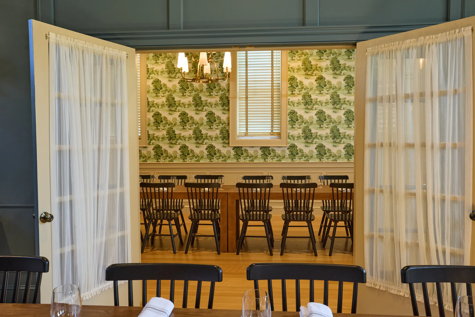 Glazed doors with sheer curtains folded open onto a room papered in green toile, a long table with black Windsor chairs inside.