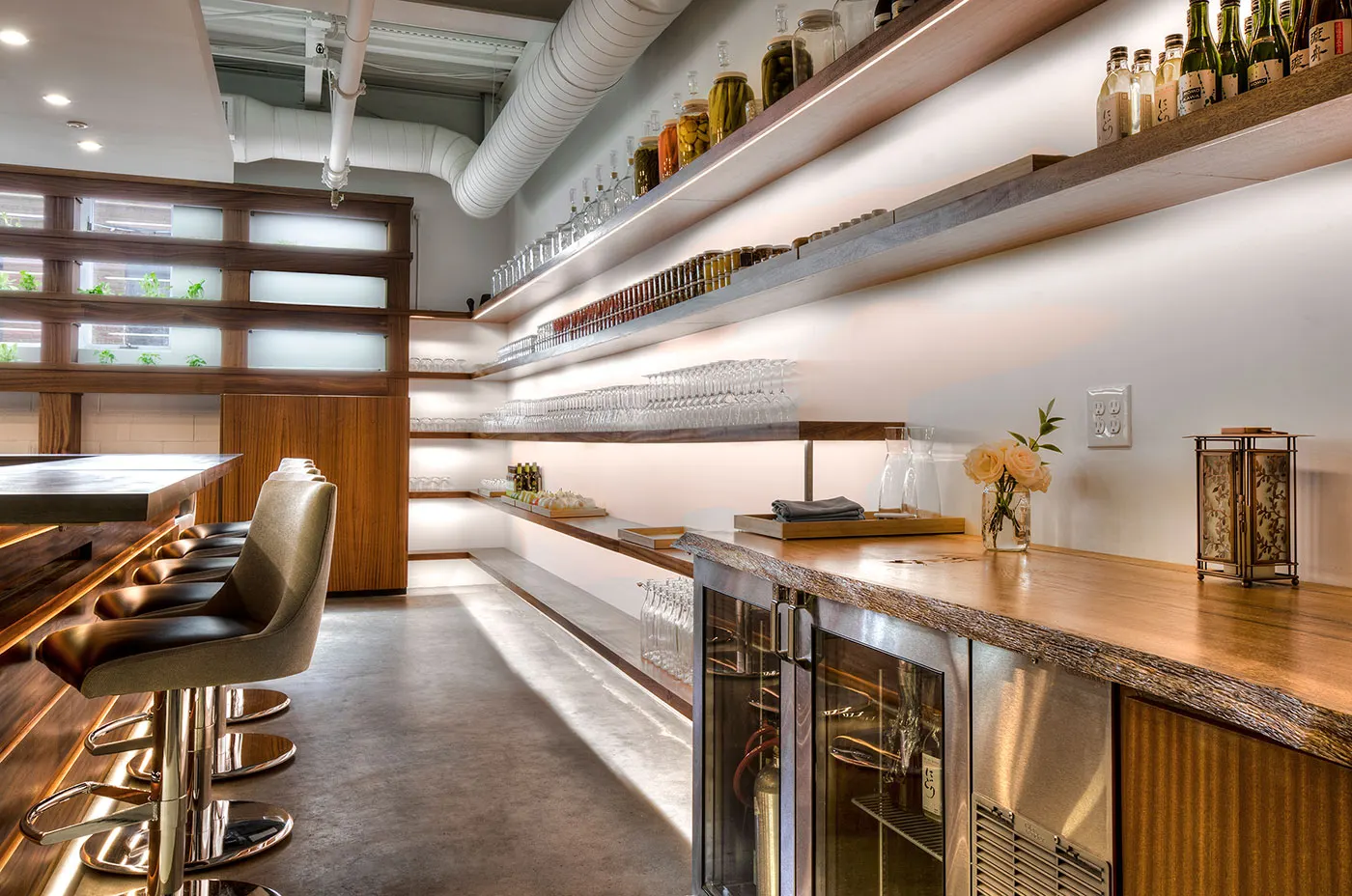 A live-edge timber counter over glass-fronted fridges, with lit shelves of jars, bottles and glassware behind.