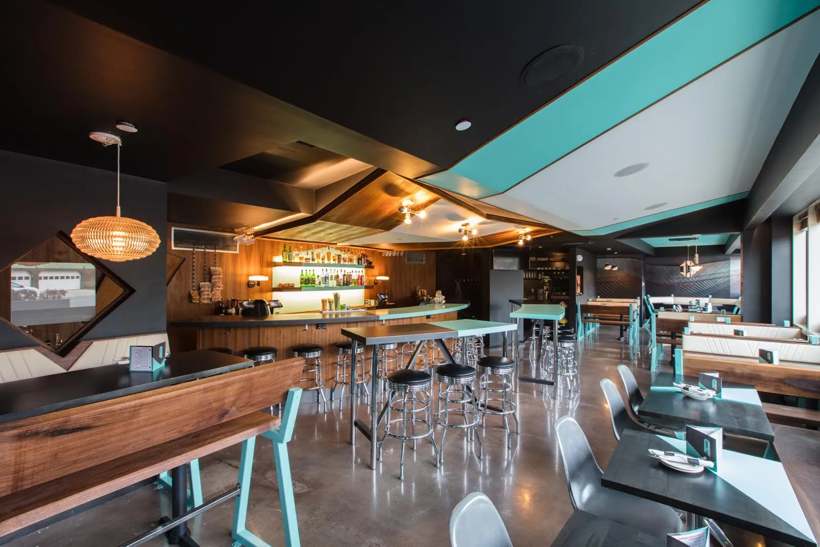 Tall tables and stools in front of a timber-lined bar, under a ceiling of teal and white panels.