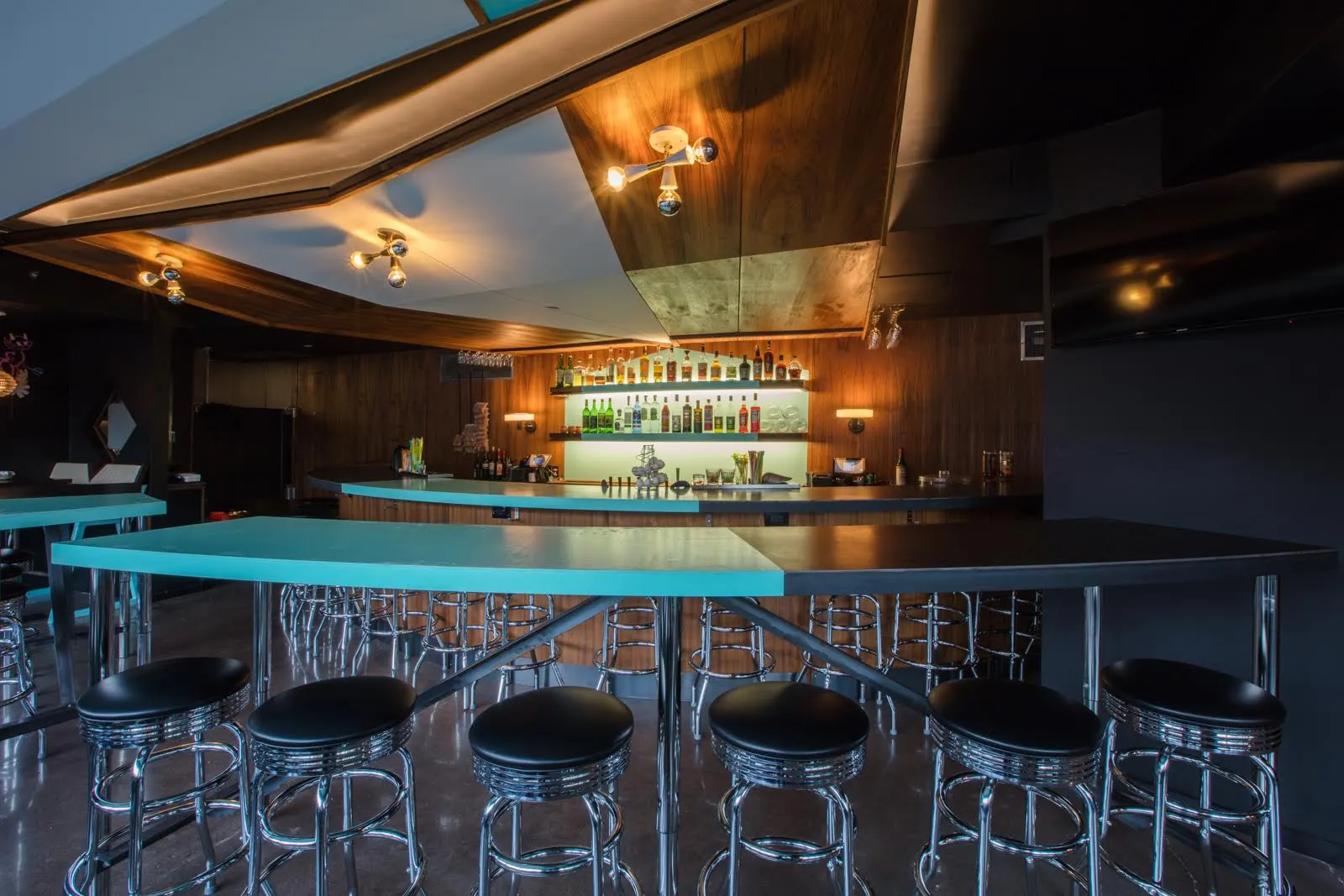 A teal counter ringed with black chrome stools, the back bar lit behind.