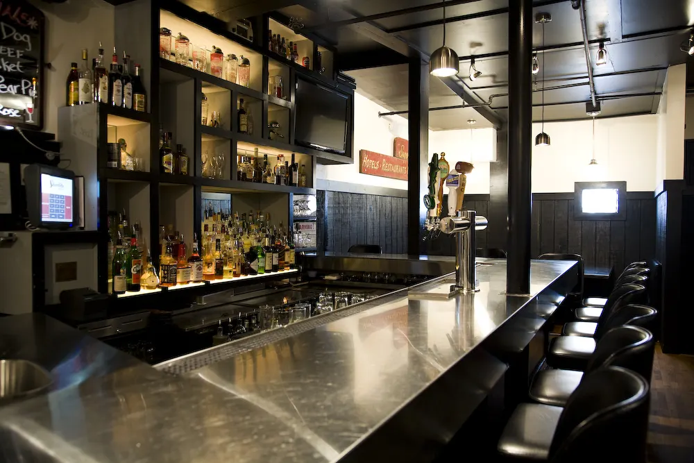 A long steel-topped bar with stools, and lit shelves of bottles behind.