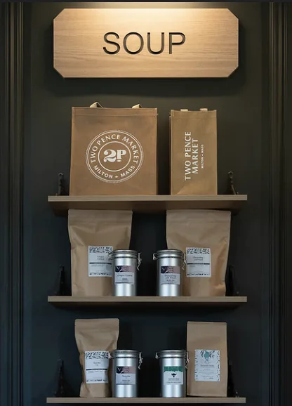 A carved SOUP sign over shelves of Two Pence Market bags and steel tins, against a dark green wall.