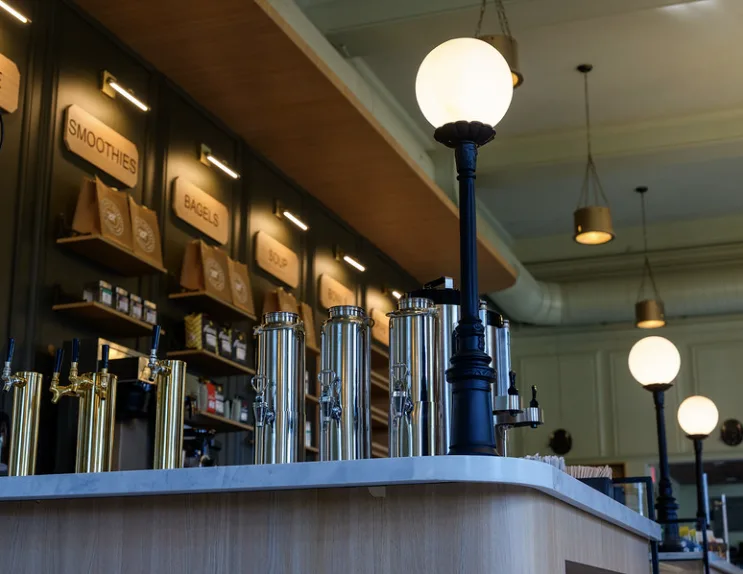 Steel urns and taps along a timber counter, under globe lamps on black posts, with a sign for each station.
