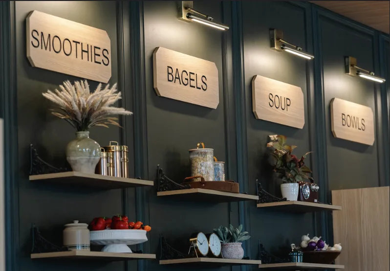 Carved signs for smoothies, bagels, soup and bowls on a dark green wall, above small display shelves.