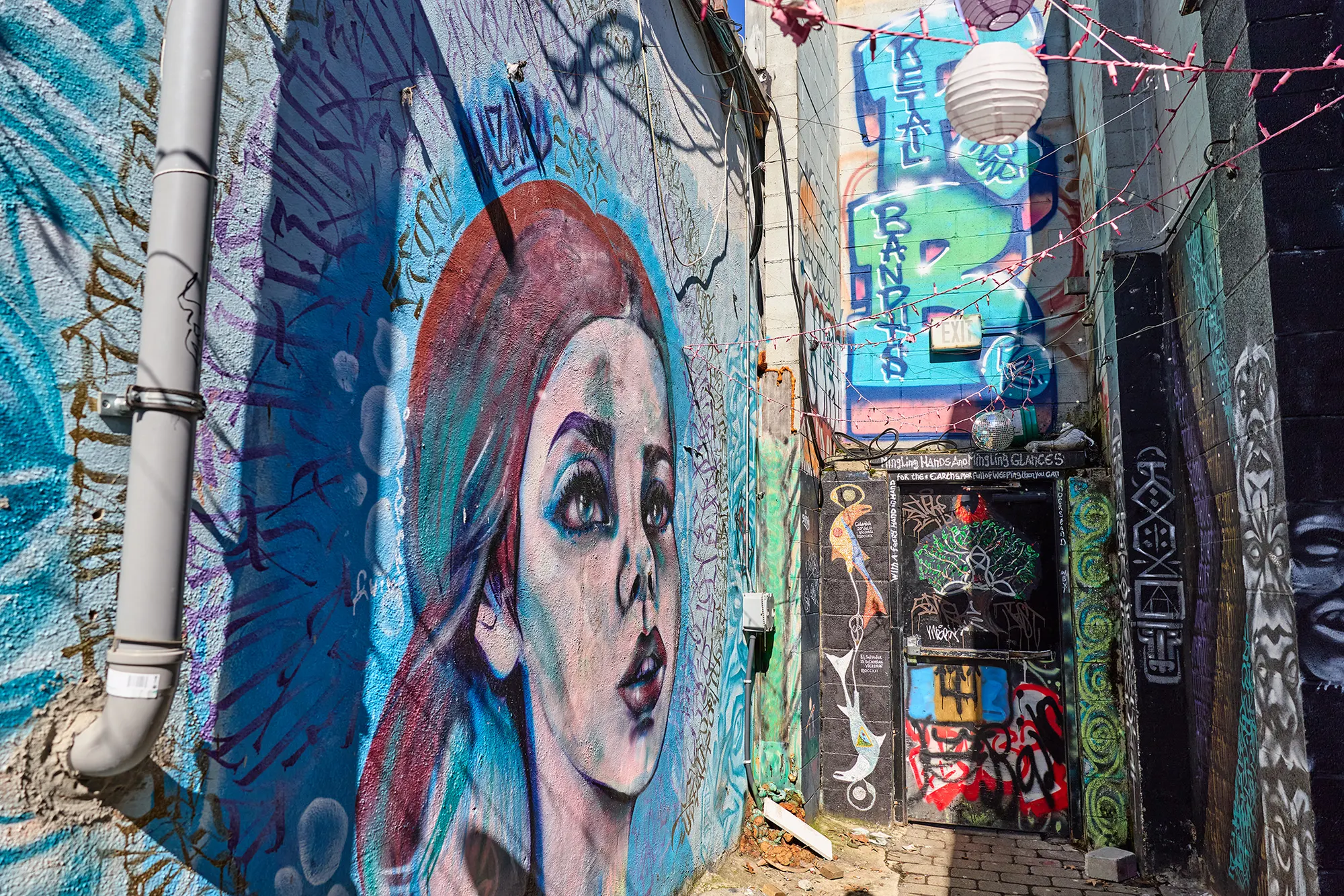 A mural of a red-haired woman in the alley, with lanterns strung above.