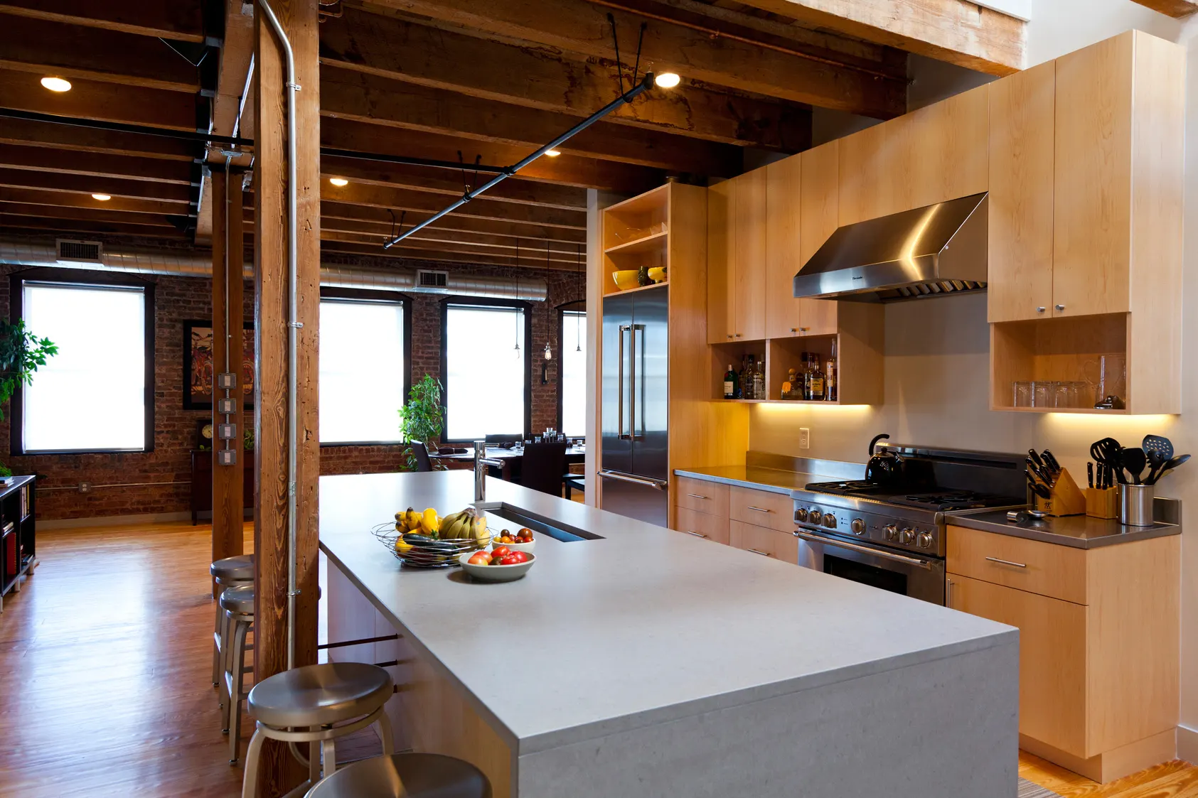 A long pale concrete island in front of maple cabinets and a stainless range, under the timber ceiling.
