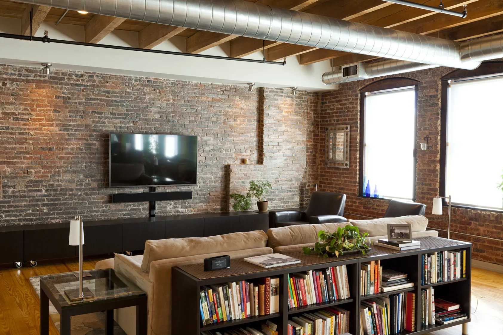 A sofa backed by a low bookcase, facing a television on a bare brick wall under exposed ductwork.