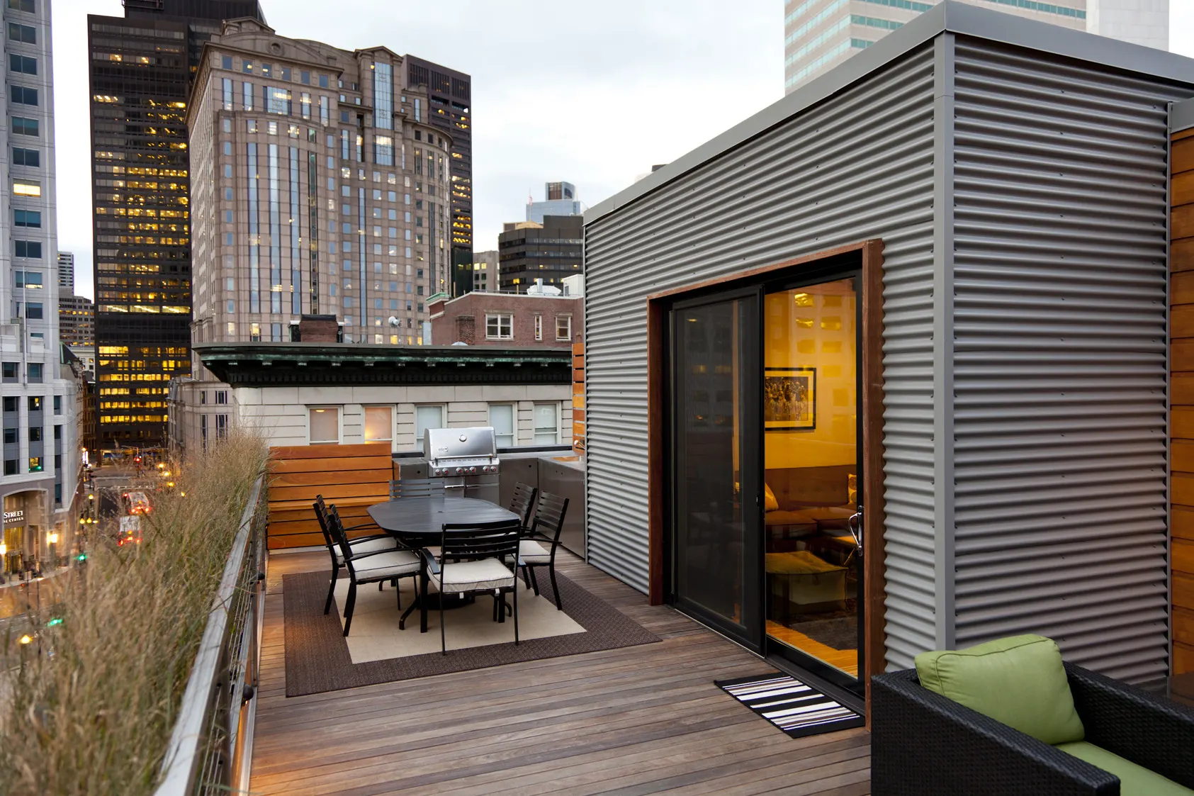 A dining table on the deck beside a penthouse clad in corrugated metal, with office towers beyond.