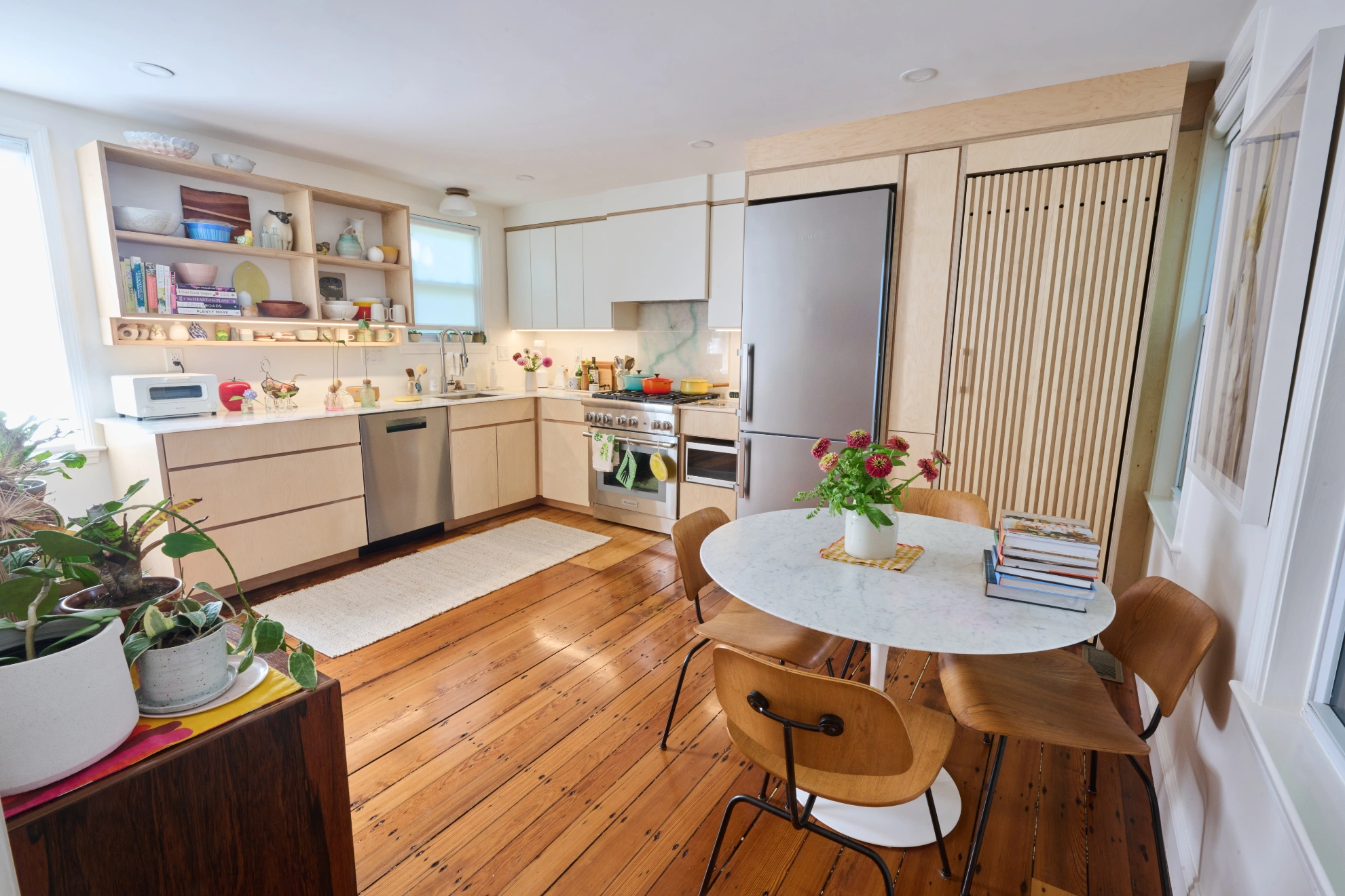 The kitchen and dining area in one view: birch cabinets and open shelves, and a round marble table on wide pine floorboards.