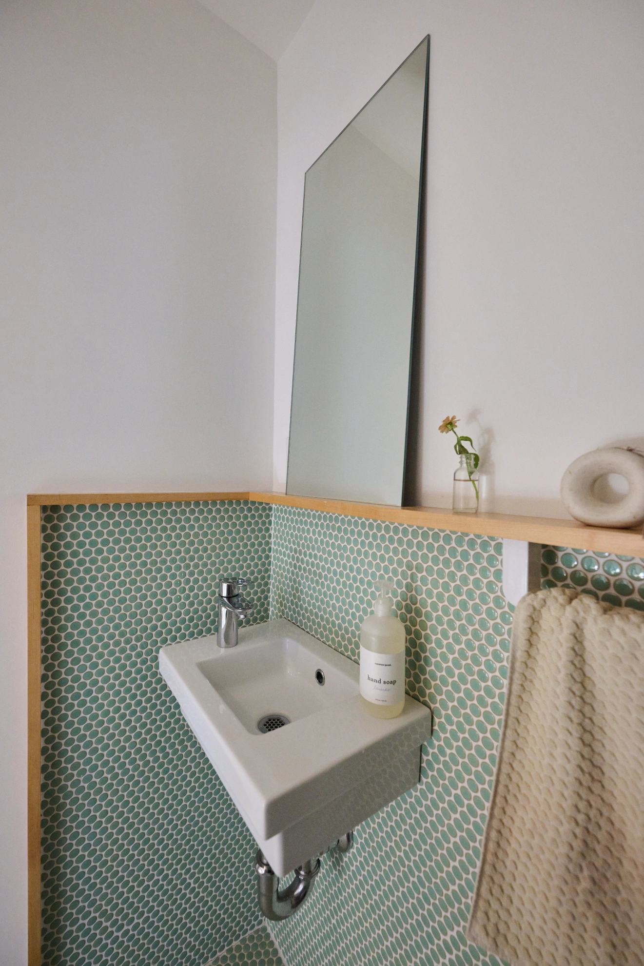 A small white basin against green penny tile under a tall mirror, with a birch shelf above it.