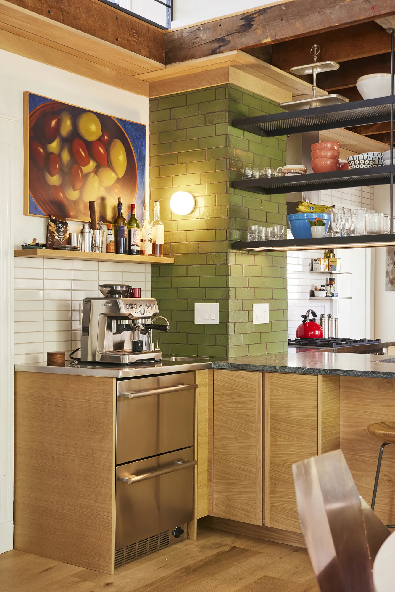 An espresso machine on timber cabinets beside a column of green tile, with steel shelves and a painting above.