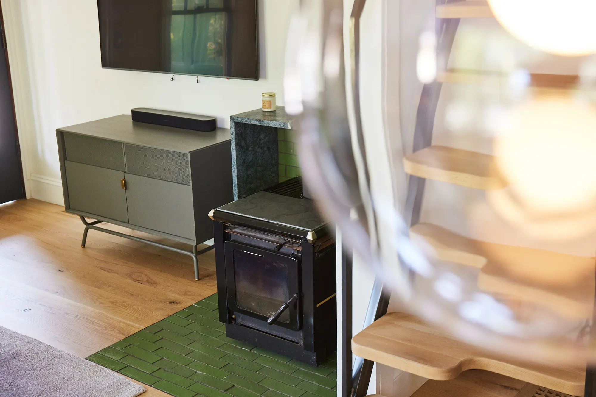 A black wood stove on green tile beside a grey media cabinet, a glass in soft focus in front.