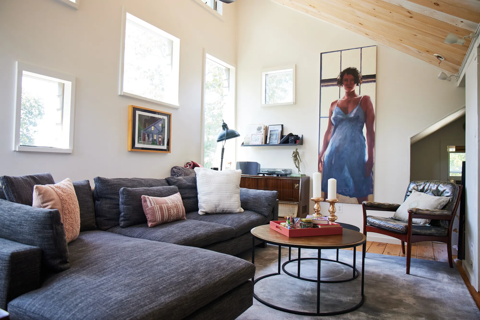 A grey sectional and a round coffee table under a pitched timber ceiling, with a tall painting of a woman on the wall.