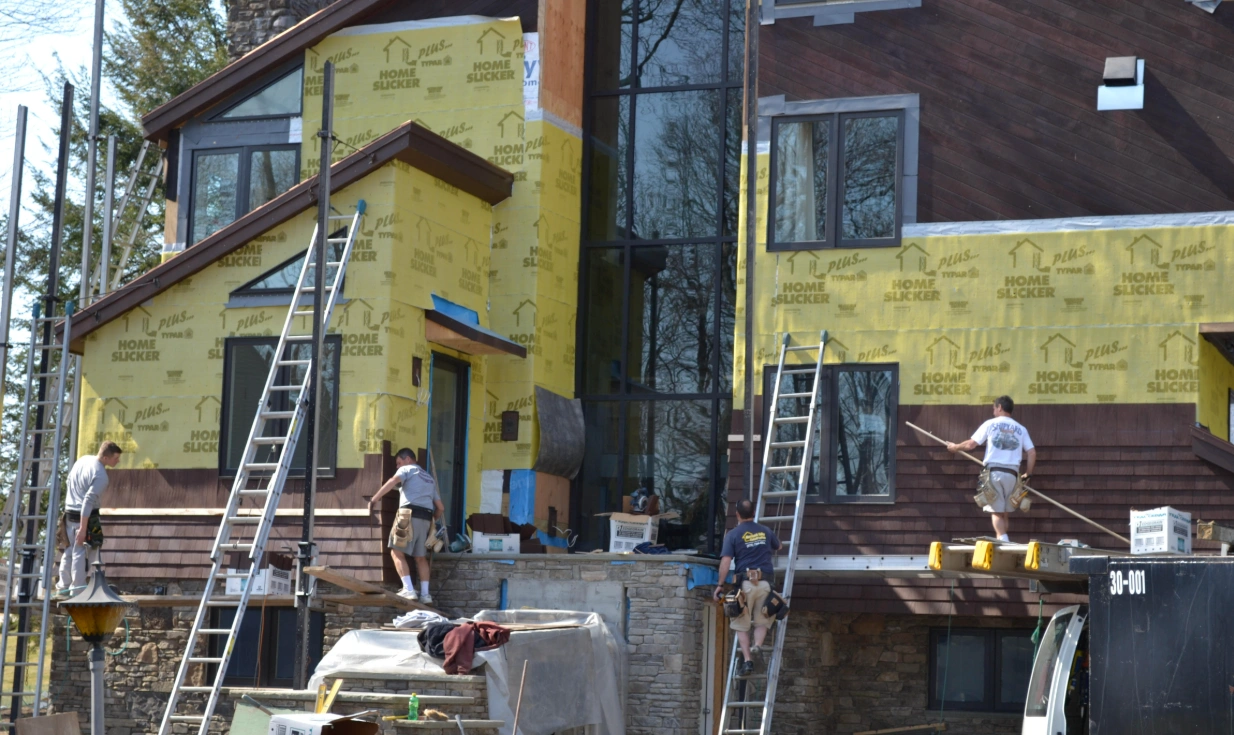 Builders on ladders against the house, part wrapped in house wrap, with the new glazed tower in place.