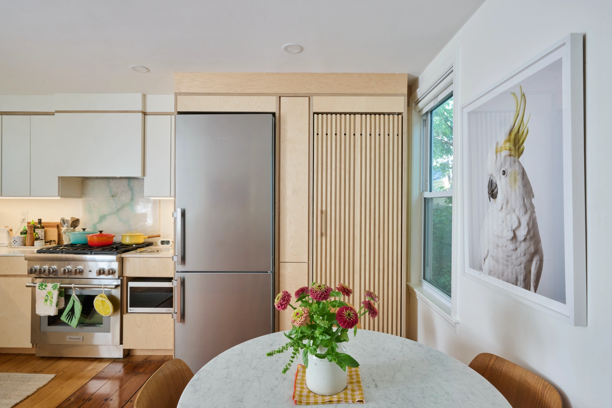 A round marble table set with flowers in front of pale birch cabinets, a steel range and refrigerator, and a framed photograph of a cockatoo.
