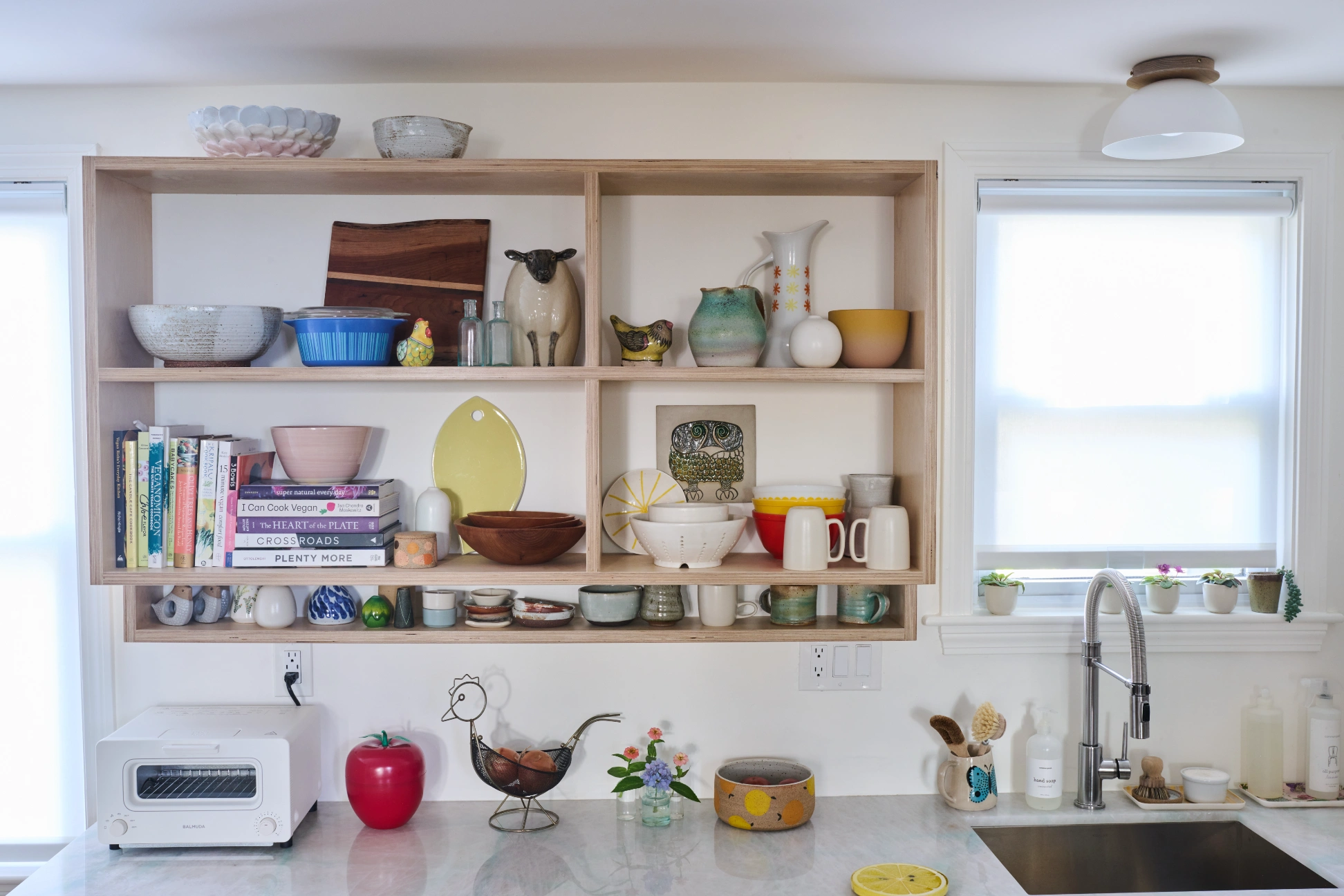 A birch shelving unit of bowls, books and pottery above the counter, between two windows.