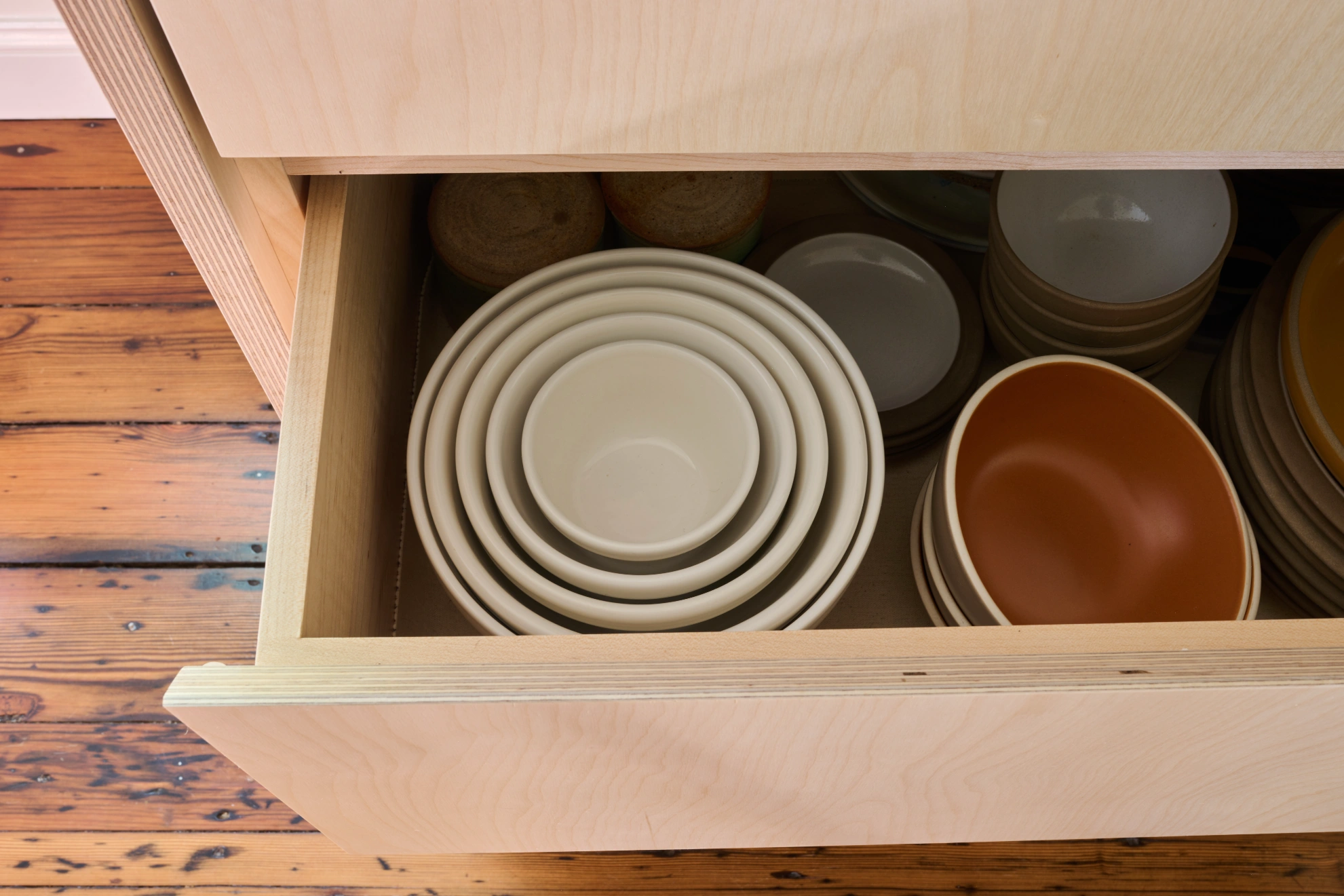 A deep birch drawer of stacked bowls and plates, pulled open over the pine floor.