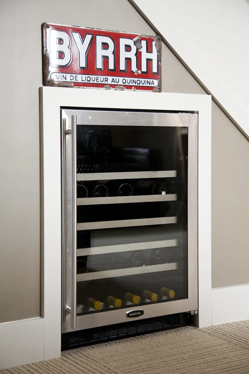 A glass-fronted wine refrigerator built in under the stair, with an old BYRRH enamel sign above.