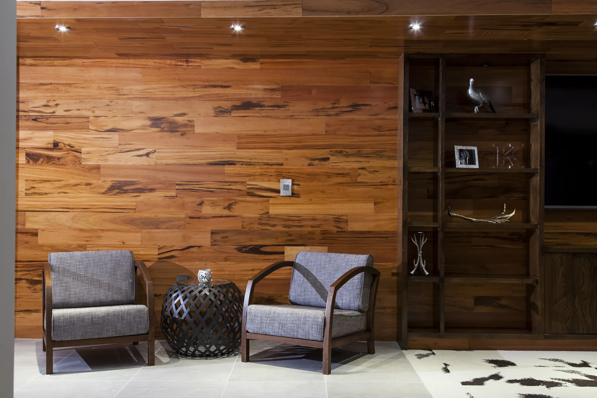 Two grey armchairs and a side table against a timber-boarded wall, beside built-in shelving.
