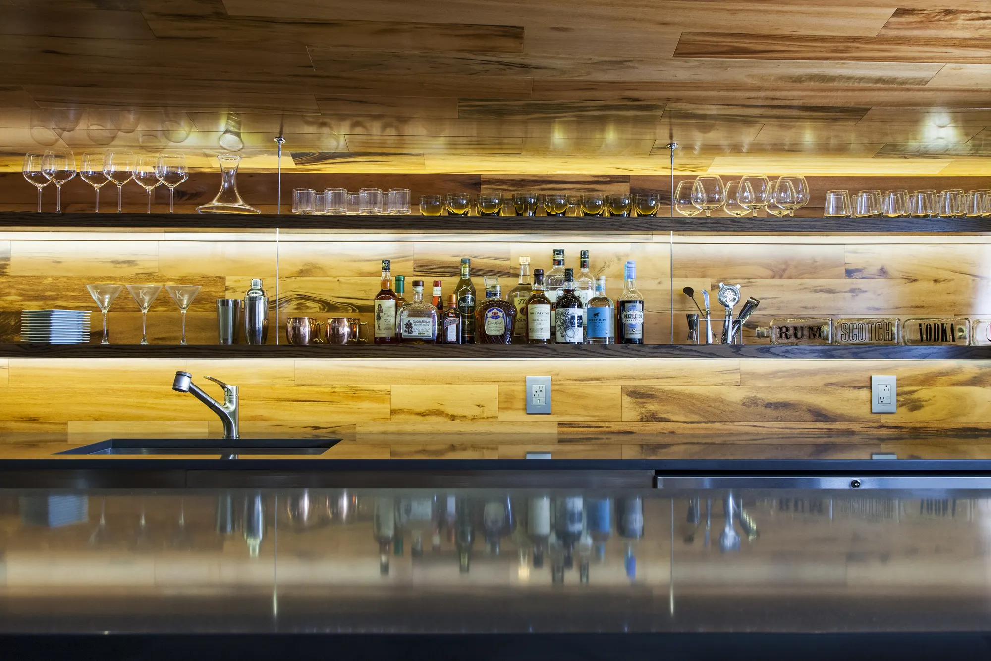 Lit shelves of glasses and bottles over a sink on a timber-clad wall, reflected in the dark bar top.